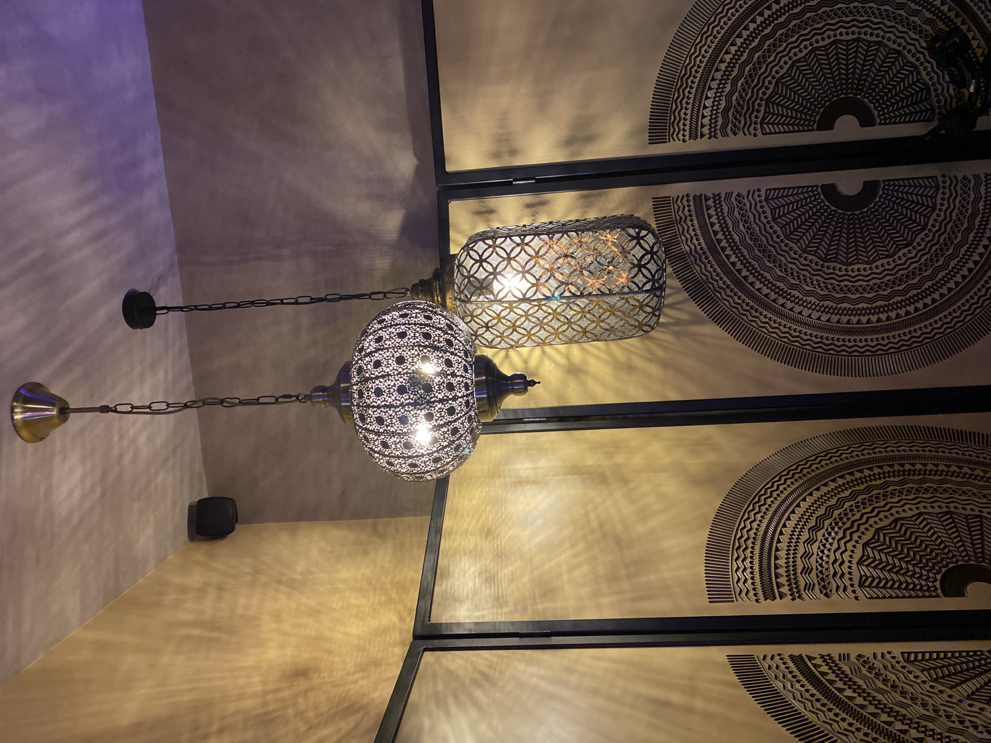 The image shows a low-angle view of two ornate metal hanging lamps, casting patterned light onto the ceiling and walls. One lamp is spherical with small perforations, and the other is rectangular with a geometric mesh pattern. The background consists of decorative wall panels with large, concentric circular designs, framed by dark, rectangular borders. The walls appear to be textured, with a color gradient from beige to light purple. There is no information about the location in the city of Izhevsk, Russia.