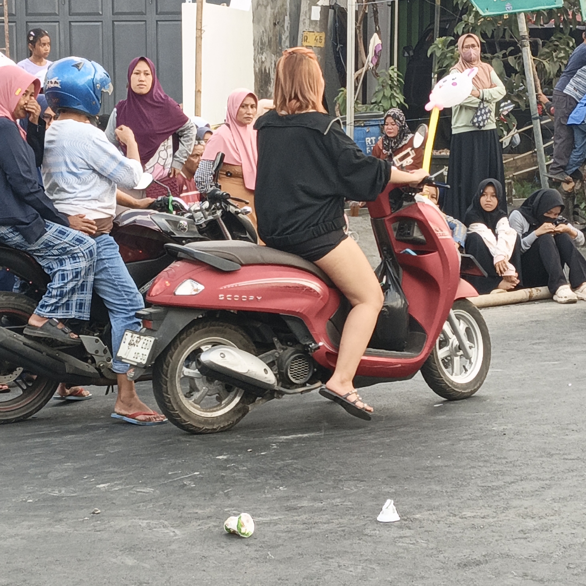 The image depicts a street scene in Randudongkal, Indonesia, with several people and two motorcycles. In the foreground, a person on a red scooter is wearing black shorts and a black top, with their legs visible. Another person in a blue helmet and patterned pants is seated on a dark motorcycle to the left, also with their legs visible. Several other individuals are standing or sitting in the background, some wearing hijabs. A woman in the middle ground is holding a pink balloon with a rabbit design. The environment appears to be an urban street with buildings and vegetation visible. The ground is paved and somewhat cluttered with debris.