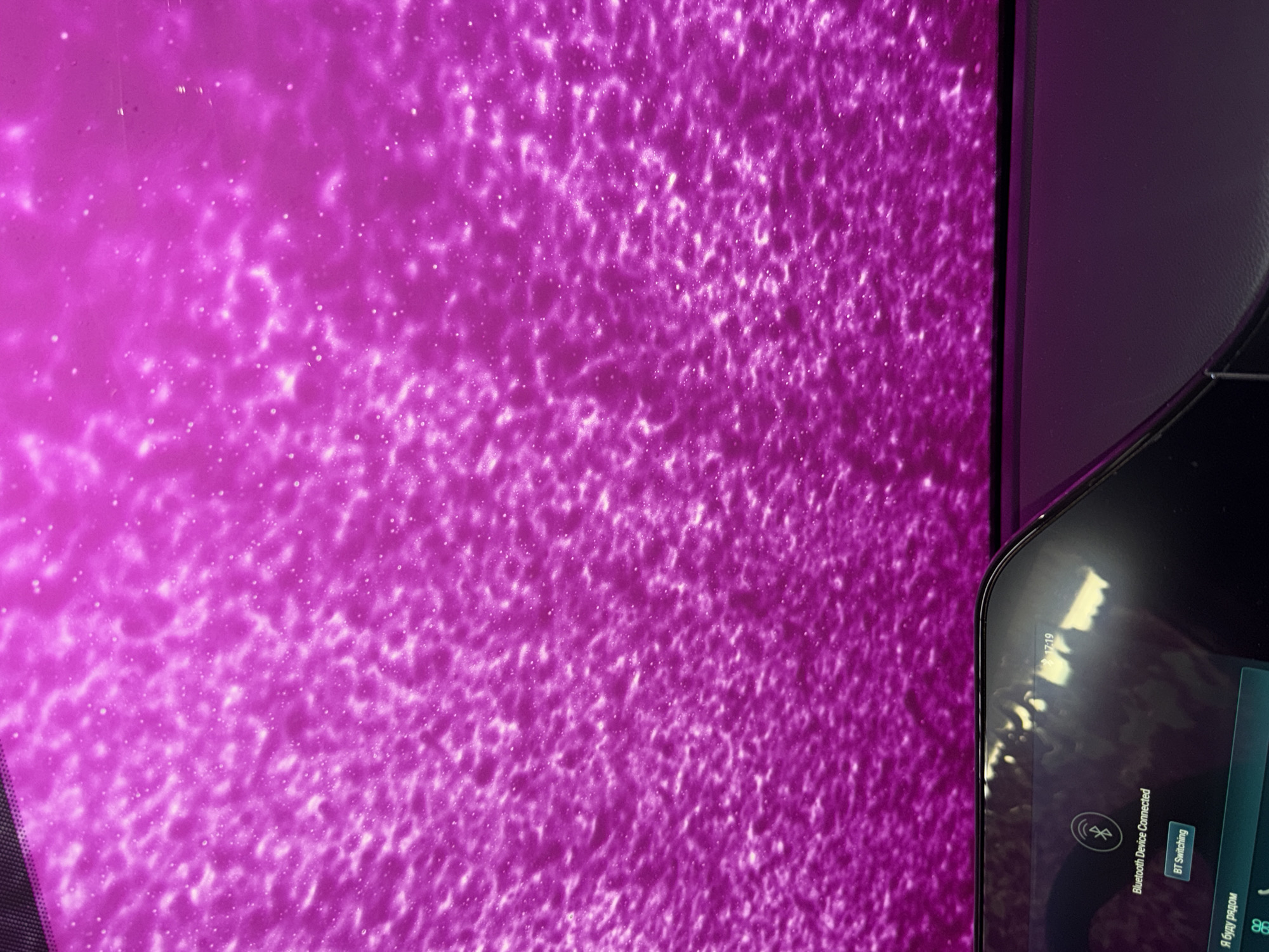 The image depicts the interior perspective from a vehicle, likely within a car wash or an environment featuring a large, illuminated magenta surface. The upper portion of the frame is dominated by a vibrant magenta expanse, exhibiting a textured, bubbly, or foamy appearance with numerous small, brightly lit, irregular spots. This surface suggests the presence of liquid and light, creating an abstract pattern. The lower portion of the image shows the top section of a modern vehicle's infotainment screen and dashboard. The screen displays a "Bluetooth Device Connected" message along with a corresponding Bluetooth icon. A "BT Switching" button is visible, and the time "17:19" is indicated in the upper right corner of the screen. Partially visible at the bottom left of the screen is Cyrillic text, "Я буду рядом" (meaning "I will be next to you" or "I will be around"), along with other interface icons. Reflections of the car's interior and the magenta exterior are visible on the screen's glossy surface. No individuals are present in the frame. The scene is located in Chaikovsky, Russia.