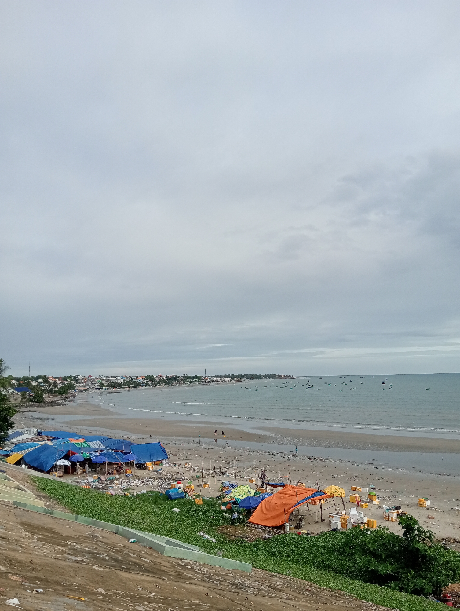 The image shows a coastal scene. In the foreground, a concrete slope is covered in vegetation. A beach is visible, covered with clusters of tents and tarps in blue, yellow, and orange. The tents seem to be used for commercial purposes, with boxes scattered around, presumably holding goods. Debris and trash are strewn across the beach. The water is calm, and small boats are scattered across the horizon. A town lines the beach in the background. The sky is overcast and gray. The location is unknown.