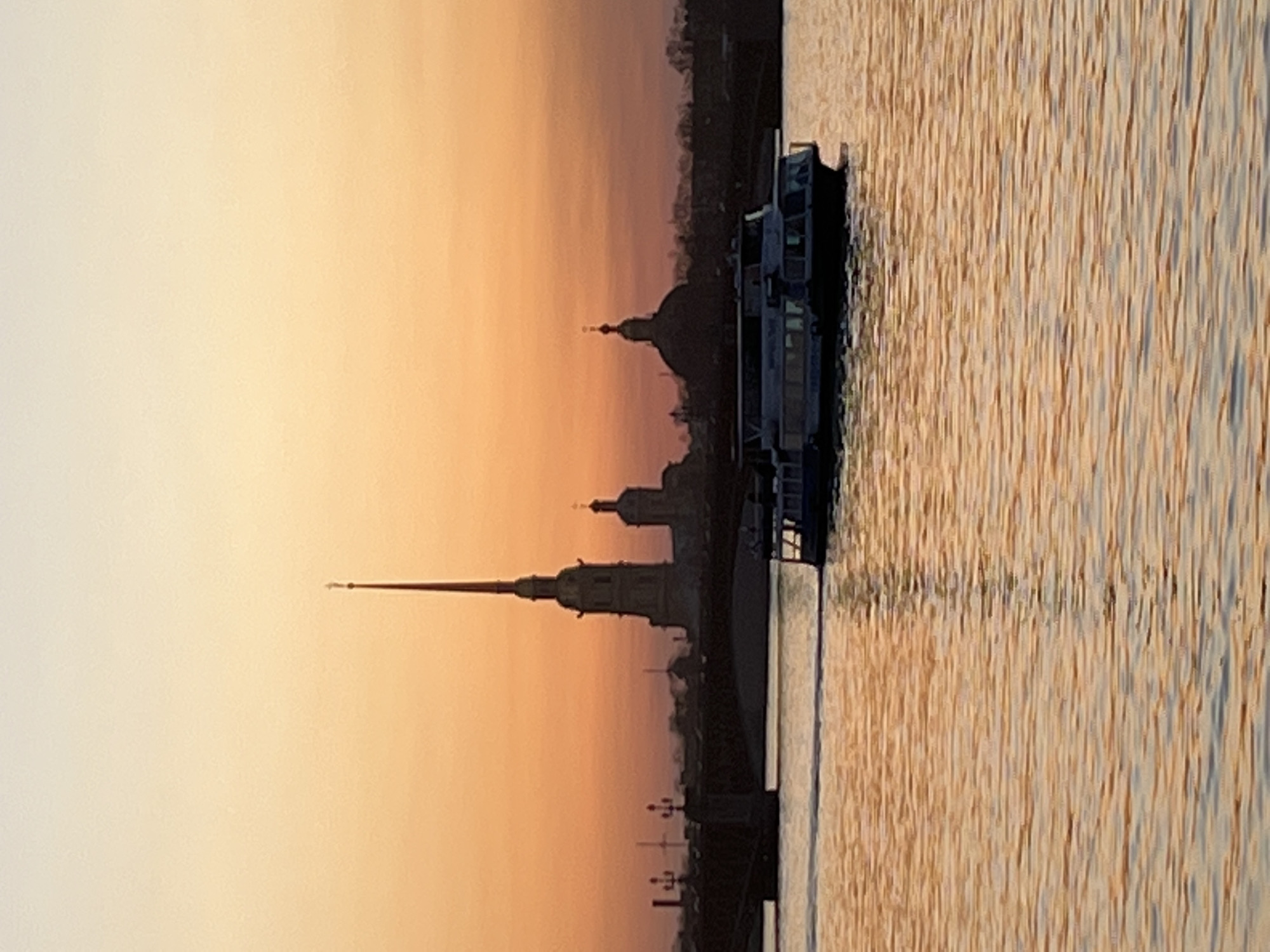 The image shows a cityscape at sunset in Saint Petersburg, Russia. The Peter and Paul Fortress is silhouetted against the orange sky. A passenger boat is floating on the water in the foreground. The water reflects the colors of the sunset.