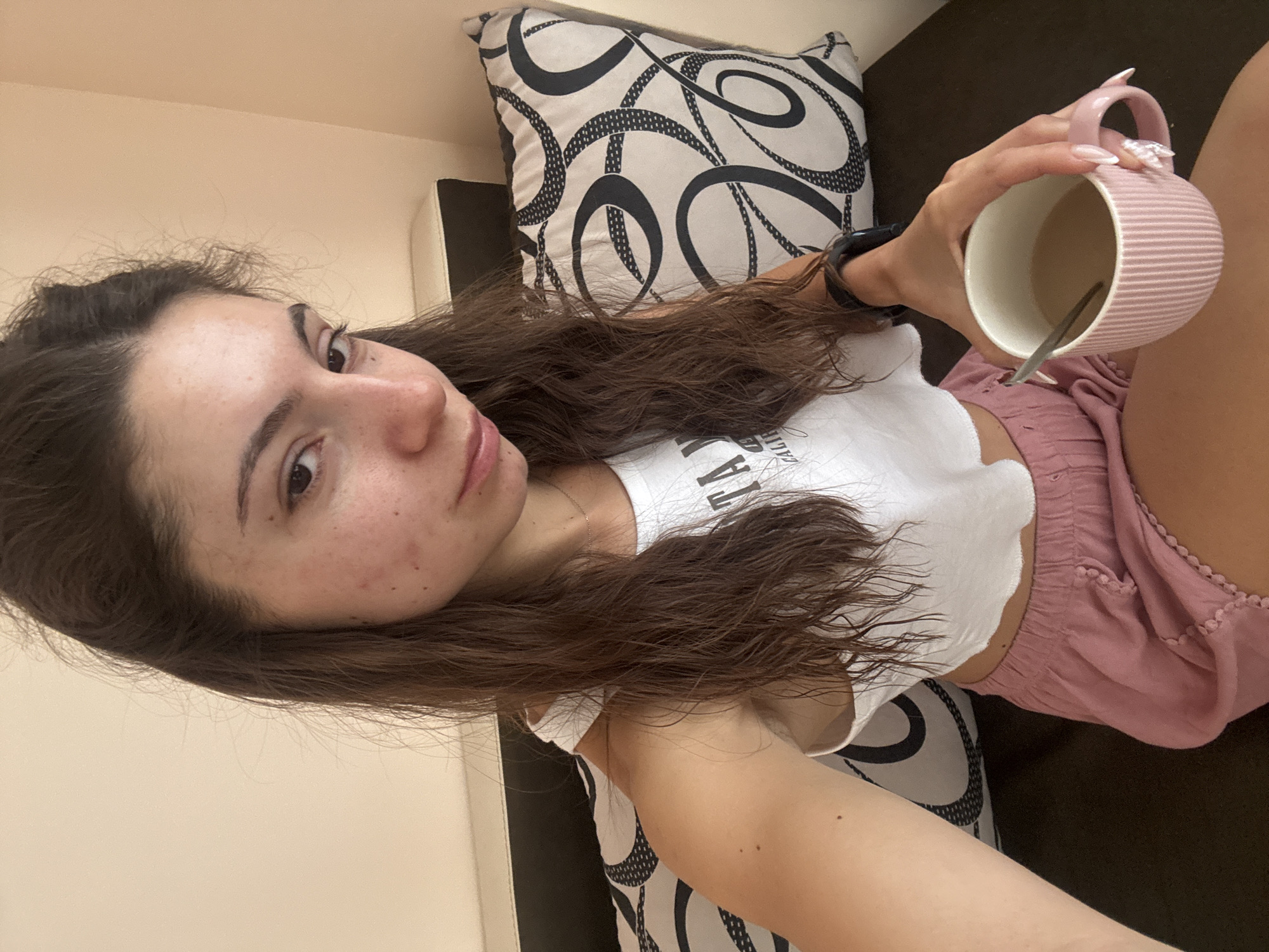 A young woman with dark brown hair and light skin is sitting on a couch. She is wearing a white crop top and pink shorts. She is holding a pink mug filled with a light brown liquid. The couch has a black cushion and a patterned pillow. The background is a plain wall. The lighting is natural, coming from the right side of the image. The overall scene suggests a casual indoor setting, possibly a home. The specific location in Varna, Bulgaria is not discernible from the image itself.
