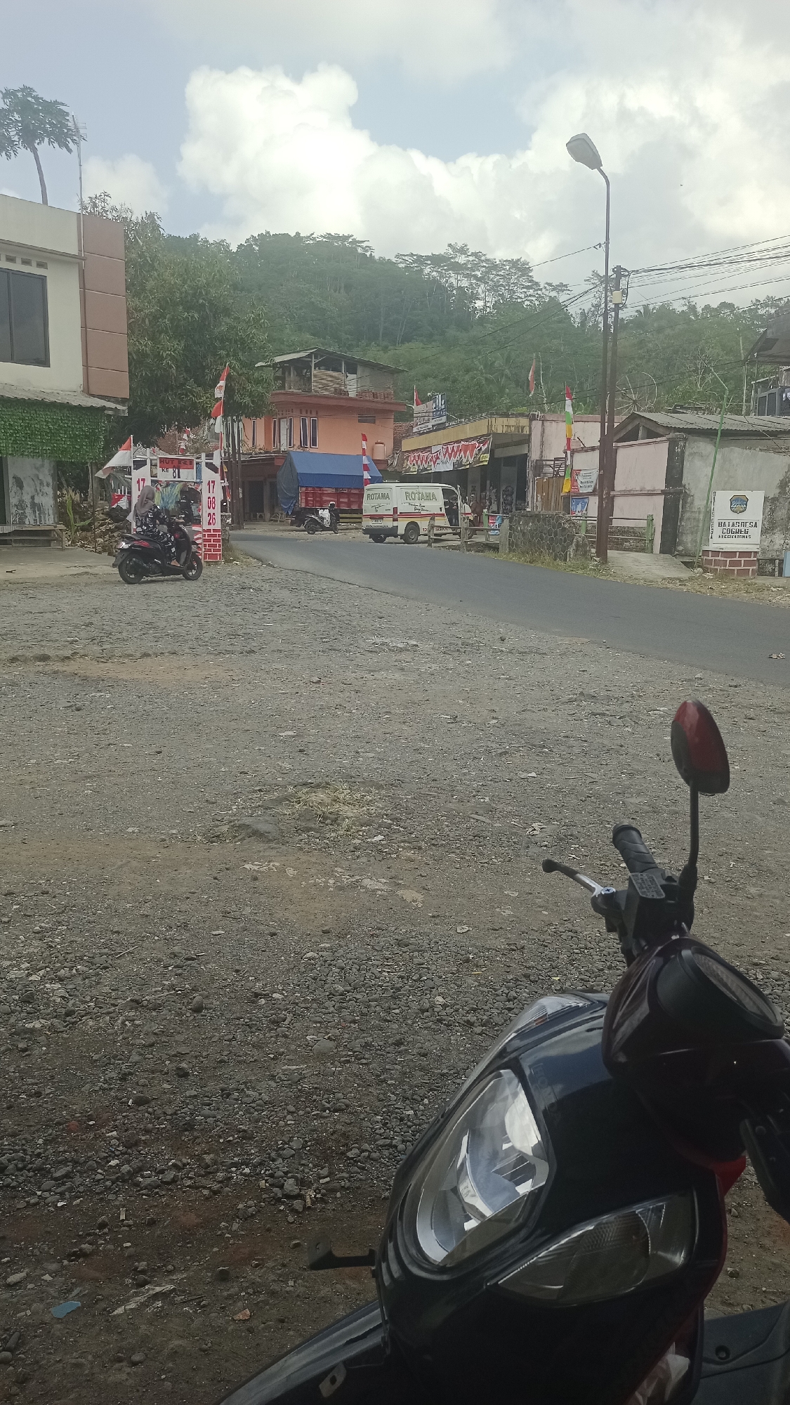 In Karyamandala, Indonesia, a street scene is captured with a focus on a motorcycle in the foreground, partially obscuring a road and various buildings. The road is paved with asphalt and bordered by a rough, gravelly shoulder. To the left, a modern building with a beige facade and dark windows stands. Further down the street, a row of shops and buildings is visible, including a pink and orange structure, and a truck with the branding "ROTAMA" and "ROTAMA LOGISTIK". A white van with similar branding is also present. Indonesian flags and banners are displayed, suggesting a local event or celebration. The background is dominated by a lush, green hillside covered in trees. The sky is partly cloudy. In the foreground, the black motorcycle's headlight, handlebars, and side mirror are clearly visible. The overall impression is of a vibrant, community-oriented street in a tropical setting.