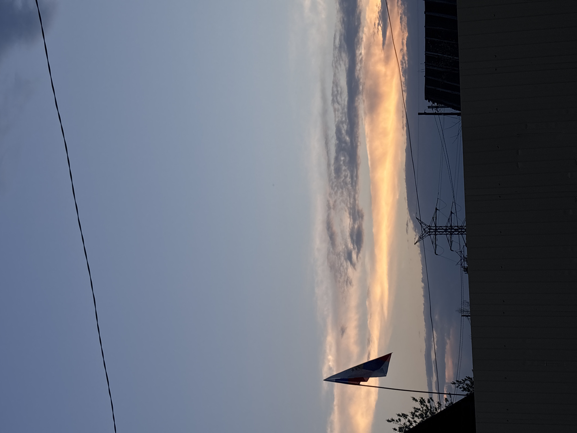 The image shows a view of the sky at dusk, likely in a residential area of Chaikovsky, Russia. A utility pole and a power line are visible on the left side. On the right, a portion of a building with vertical siding and a metal roof is seen. An electrical transmission tower is near the building. A flag of Russia is mounted on a pole. The sky is filled with clouds tinted with orange hues from the setting sun.