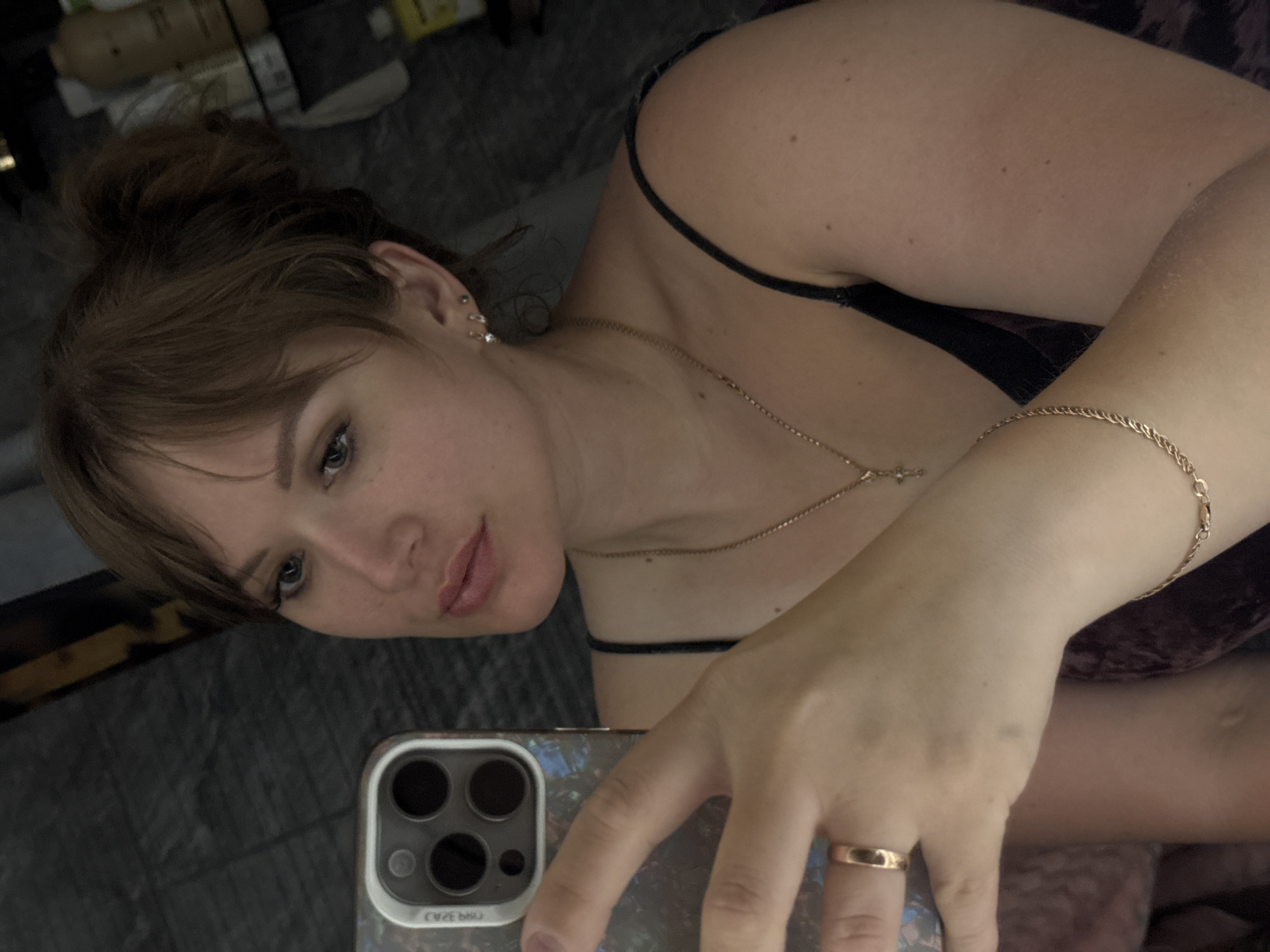 A young woman is taking a selfie indoors. She has light skin, brown hair pulled into a messy bun, and is wearing a black camisole. She has a gold necklace with a cross pendant, a gold bracelet, and a gold ring on her ring finger. She is holding a smartphone with a triple-lens camera. The background is blurry and cluttered with various items. The location is not identifiable as Chaikovsky, Russia.