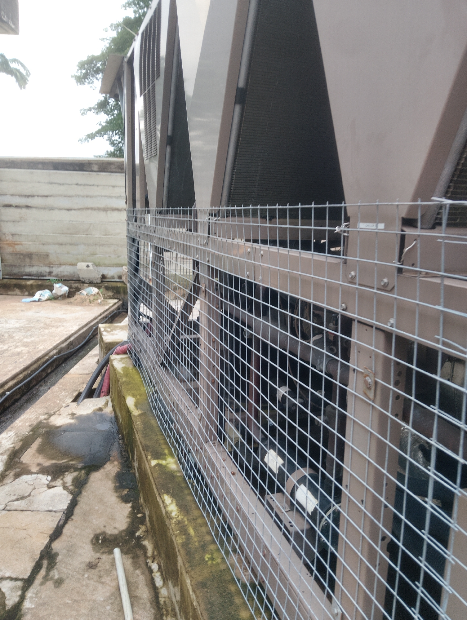 This image depicts the exterior of a large industrial cooling unit, likely an air conditioning or refrigeration system, situated outdoors. The unit is encased in a brown metal structure with visible ventilation fins and a protective wire mesh cage surrounding it. The scene appears to be in a somewhat neglected urban or industrial setting, with weathered concrete surfaces, some debris, and overgrown moss on a low wall in the foreground. The background shows a concrete wall and some trees, suggesting an outdoor rooftop or ground-level installation. The overall impression is one of functional industrial equipment in a typical outdoor environment. The question asks to include the location in the city of Onibudo, Nigeria, but there are no visible elements in the image that can confirm or suggest this specific location.