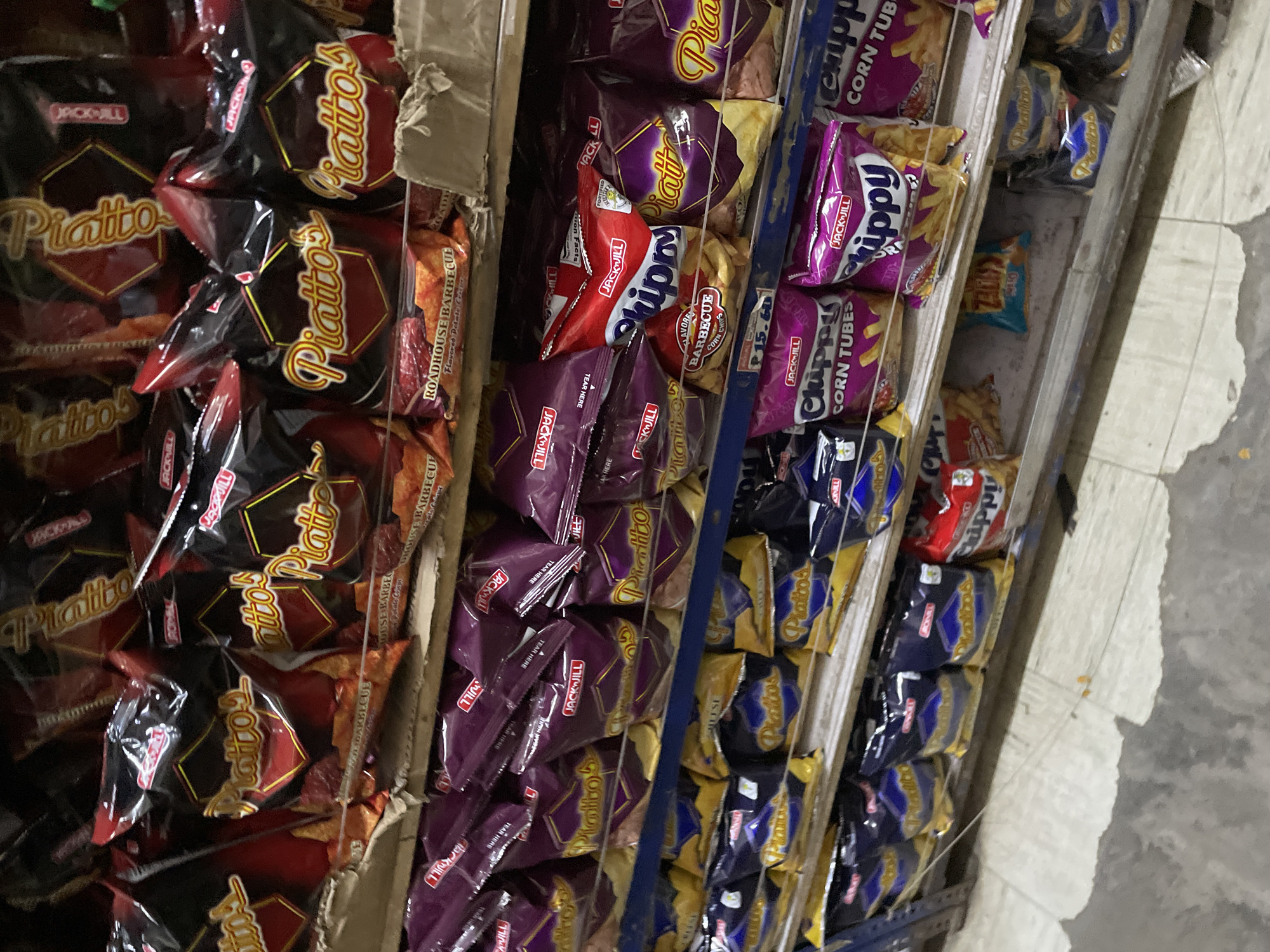 This image depicts a multi-tiered shelving unit within a retail establishment, presumed to be in Davao City, Philippines. The shelves are densely stocked with various snack bags, predominantly from the Jack 'n Jill brand.
The uppermost visible shelf displays multiple black and red bags of "Piatto's ROADHOUSE BARBECUE Flavored Potato Chips." The shelf directly below it also features numerous "Piatto's" potato chips in similar black and red packaging, along with some purple-packaged "Piatto's." The mid-level shelf holds purple and yellow "Piatto's" bags, interspersed with red and white bags of "Jack 'n Jill Skippy Barbecue Flavored Corn Chips." A price tag labeled "P 15.60" is attached to the front edge of this shelf. Below, blue and yellow "Piatto's" bags are arranged alongside purple bags of "Jack 'n Jill Chippy CORN TUBES." A small red bag of "Jack 'n Jill Skippy Corn Chips" is also visible on this level. The lowest visible shelf continues to display "Piatto's" and "Chippy" products, with an additional small blue-green bag present.
The shelving structure comprises metal supports with wooden planks, some of which appear worn. The floor is a light-colored concrete surface, exhibiting various patches of dirt, scuff marks, and areas of discoloration. No individuals are depicted in the scene.