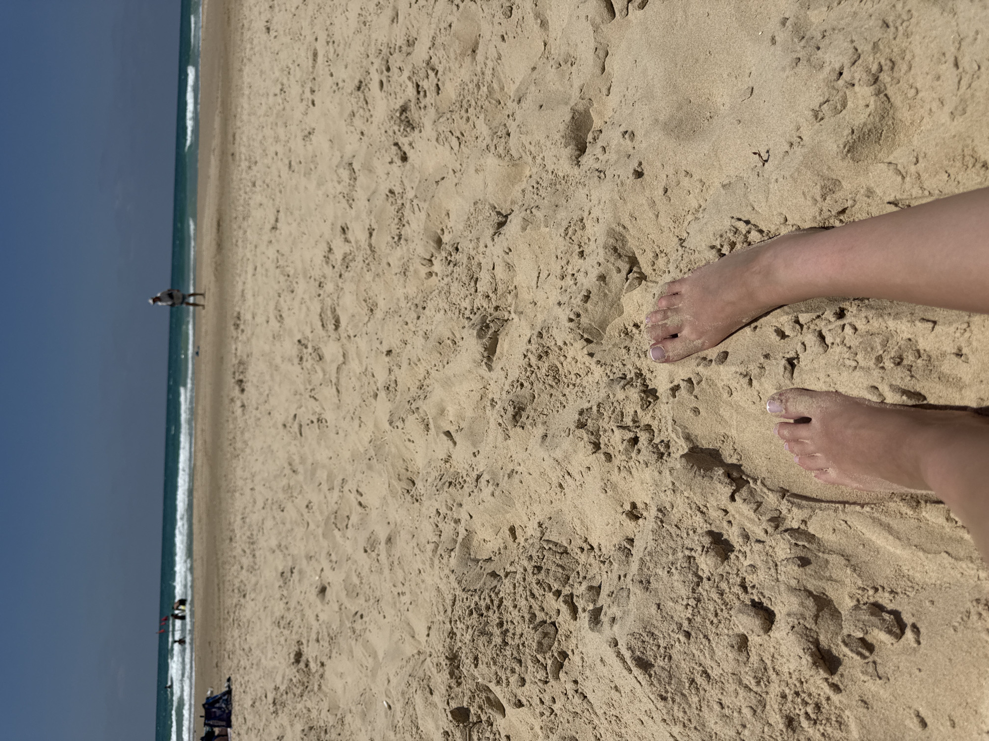 The media file shows a beach scene from a low angle, focusing on the sandy ground and a person's feet. The sand is light brown and textured, with visible footprints and indentations. Two bare feet, belonging to the same person, are prominent in the foreground, placed slightly apart on the sand. The feet appear to be clean and well-maintained, with visible toenails.
In the background, a strip of ocean is visible with gentle waves rolling onto the shore. The water is a clear blue-green color. Beyond the ocean, a person can be seen walking along the shoreline, appearing as a small, silhouetted figure. Further down the beach, a few other people are scattered, and some beach equipment, possibly chairs and an umbrella, are visible. The sky is a clear, pale blue, suggesting a bright, sunny day. The scene evokes a sense of relaxation and leisure, typical of a beach environment.
The location is Bat Yam, Israel. There is no visible text in the media file. The time of day appears to be daytime, likely late morning or afternoon, given the brightness and the clear sky. The weather seems pleasant and sunny.