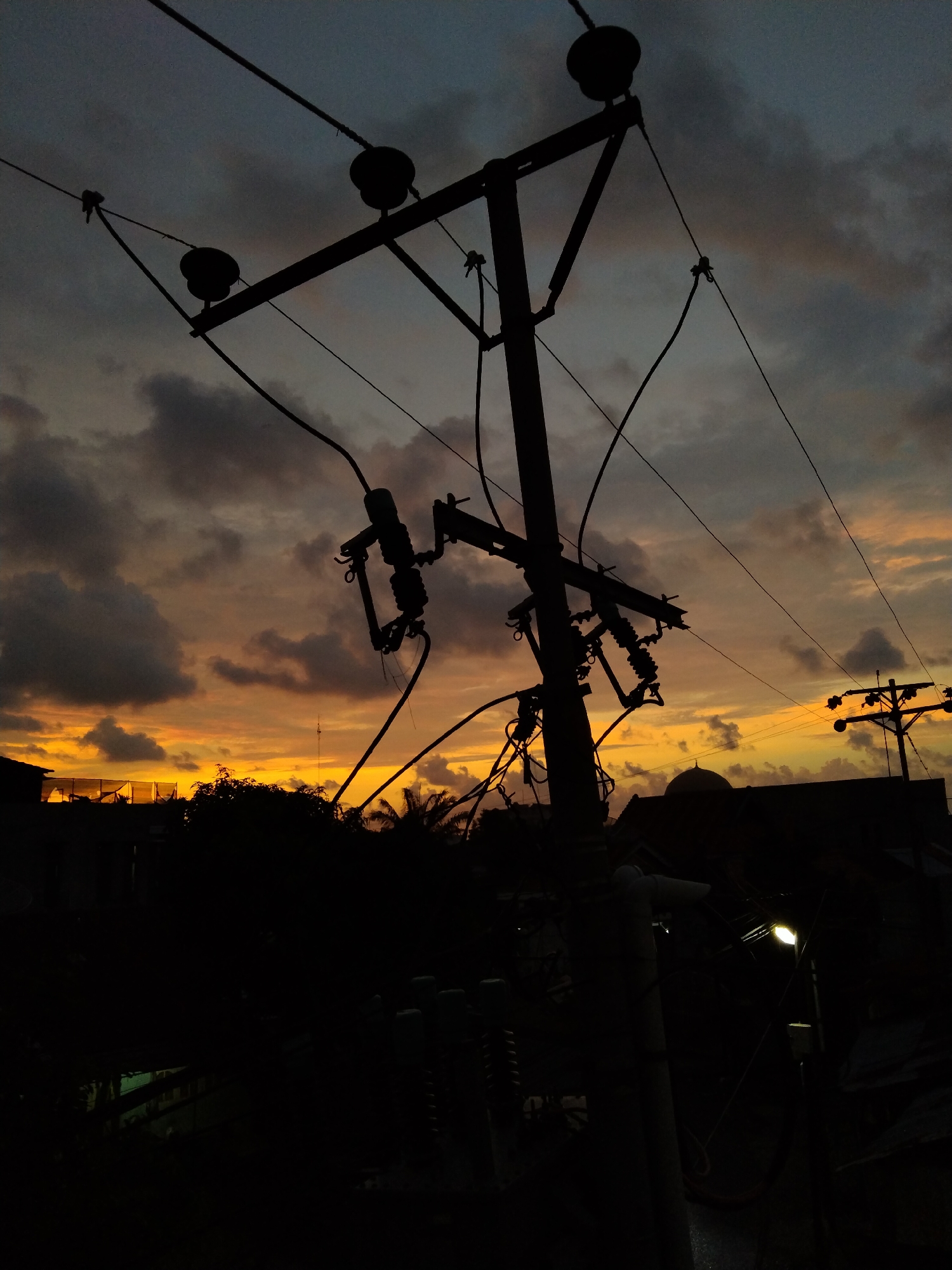 This image captures an outdoor scene at sunset in Bireuenmeunasahdayah, Indonesia, characterized by prominent silhouettes against a colorful sky. The central subject is a large utility pole, heavily laden with multiple cross arms, insulators, and a dense network of electrical or telecommunication wires, all rendered in dark silhouette against the sky.
The sky itself displays a dramatic gradient, transitioning from dark, moody grey clouds at the top to vibrant shades of orange, yellow, and red closer to the horizon, indicative of a vivid sunset. Scattered clouds are visible throughout the sky.
In the mid-ground, silhouetted shapes of other, smaller utility poles, various buildings, and dense foliage, including what appear to be palm trees, can be seen. A distinctive domed roof, possibly belonging to a mosque, is visible among the buildings on the right side. On the far left, a structure with a visible mesh or net covering is also silhouetted. The lower portion of the image is plunged into deep shadow, rendering details almost invisible, save for a single, glowing outdoor lamp or street light on the right, which casts a faint, warm light.
The scene conveys a peaceful transition from day to night in an urban or semi-urban setting. There are no people visible, and no specific activity is taking place. No visible text is present in the image.