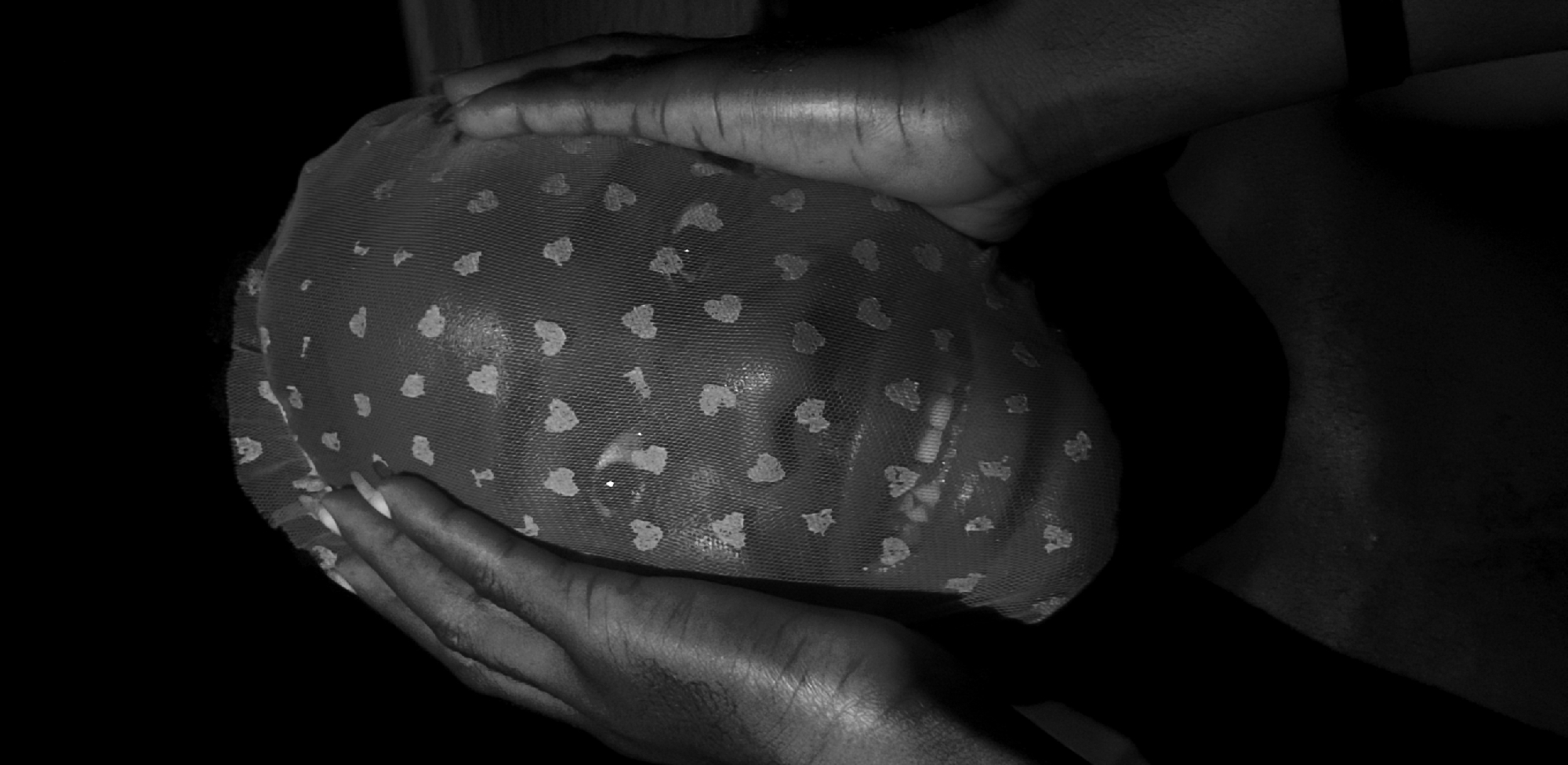 A close-up, black and white photograph shows two dark-skinned hands holding a large, round object covered in a patterned mesh. The mesh has regularly spaced heart shapes on it. Through the mesh, a human face is faintly visible, suggesting the object may be a covering over the person's head. The background is entirely black, making it impossible to determine the environment. No evidence suggests this image was taken in Narayi, Nigeria.