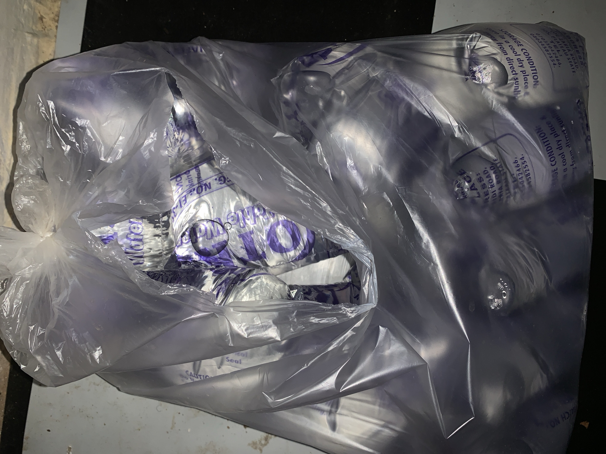 A large, clear plastic bag, appearing full and crinkled, is the central subject of the image. The bag contains numerous smaller, individual sachets or pouches made of clear plastic, which hold a transparent liquid, most likely water. These inner sachets feature purple text and designs. The large bag is resting on a light-colored, possibly concrete or tiled, surface, with a dark, indistinct background.
The image captures a static scene of packaged liquid intended for consumption. No activity is taking place; it appears to be a stored or prepared batch of goods.
Visible text on the outer bag or internal packaging provides significant details. Key phrases include "STORAGE CONDITION: Store in a cool dry place & keep away from direct sunlight" and "CAUTION Seal." Address details are clearly legible: "ZARIA ROAD," and "PLATEAU STATE." Accompanying telephone numbers are "07035042466" and "08034582554." Other partial text includes "G. NO.: E1-3," "Water," and fragments of numbers like "1582554" and "5042466." The packaging style, featuring individual sachets within a larger bag, is typical for "sachet water" or "pure water" commonly sold in Nigeria. The explicit mention of Zaria Road and Plateau State directly ties the image to a Nigerian context. The lighting is artificial, casting reflections on the plastic, suggesting an indoor setting. No people are visible, and no specific time of day or weather can be determined.