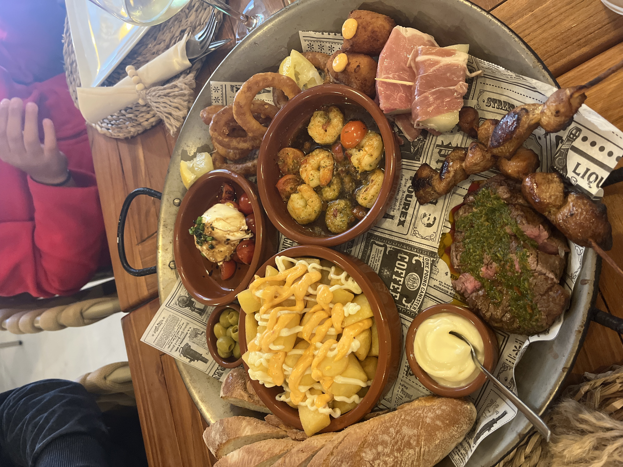 Here is a description of the image:
The image shows a high-angle view of a large, round metal tray filled with a variety of tapas-style dishes. The tray sits on a wooden table with a decorative woven mat visible beneath it. A child's hand clad in a red sleeve is partially visible at the upper left corner of the image, suggesting the food is being shared or enjoyed at a table.
The tray contains several small brown bowls and individual servings of food items arranged on newspaper-style paper with vintage advertisements and graphics printed on it. The dishes include:
* Fried potatoes drizzled with sauces.
* Garlic Shrimp.
* Burrata with tomatoes.
* Skewered meat.
* Croquettes.
* Prosciutto wrapped around mozzarella balls.
* A steak cut and topped with herbs.
* A bowl of mayonnaise with a spoon.
* A side dish of olives.
* A portion of bread.
* Onion rings
The composition of the tray suggests a shared meal or tasting experience at a restaurant, likely in Xàbia / Jávea, Spain, a popular tourist destination.