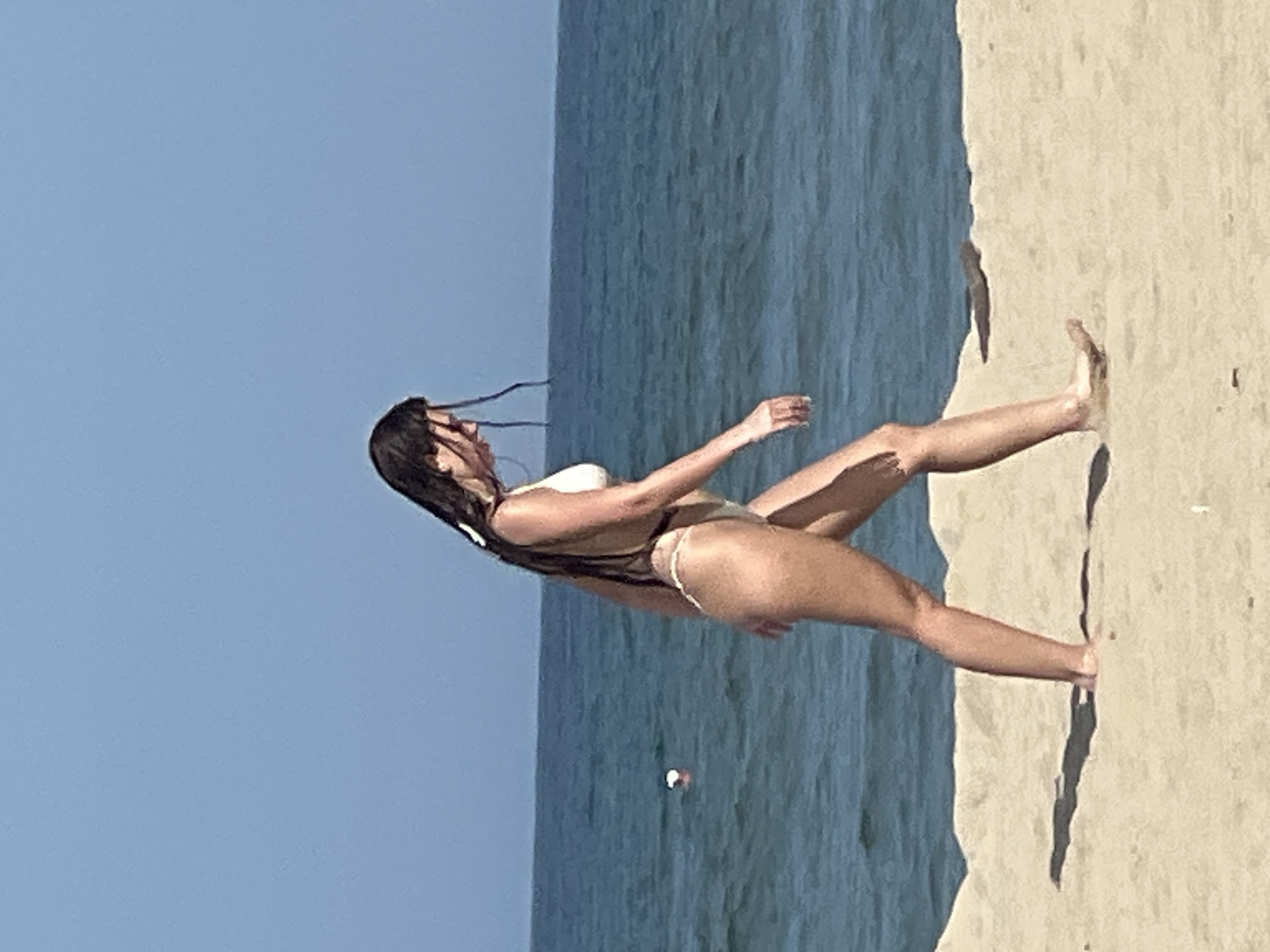 A woman in a white bikini walks along the sandy shore of a beach in Da Nang, Vietnam. The ocean, a vibrant blue, stretches out behind her under a clear sky. Her dark hair is wet and trails down her back. She is walking away from the camera, her body angled slightly towards the water. The sand is light brown and textured, with a few small shadows cast by the sun. A white buoy with an orange top is visible in the water to the left of the woman. The overall scene depicts a peaceful moment at the beach.