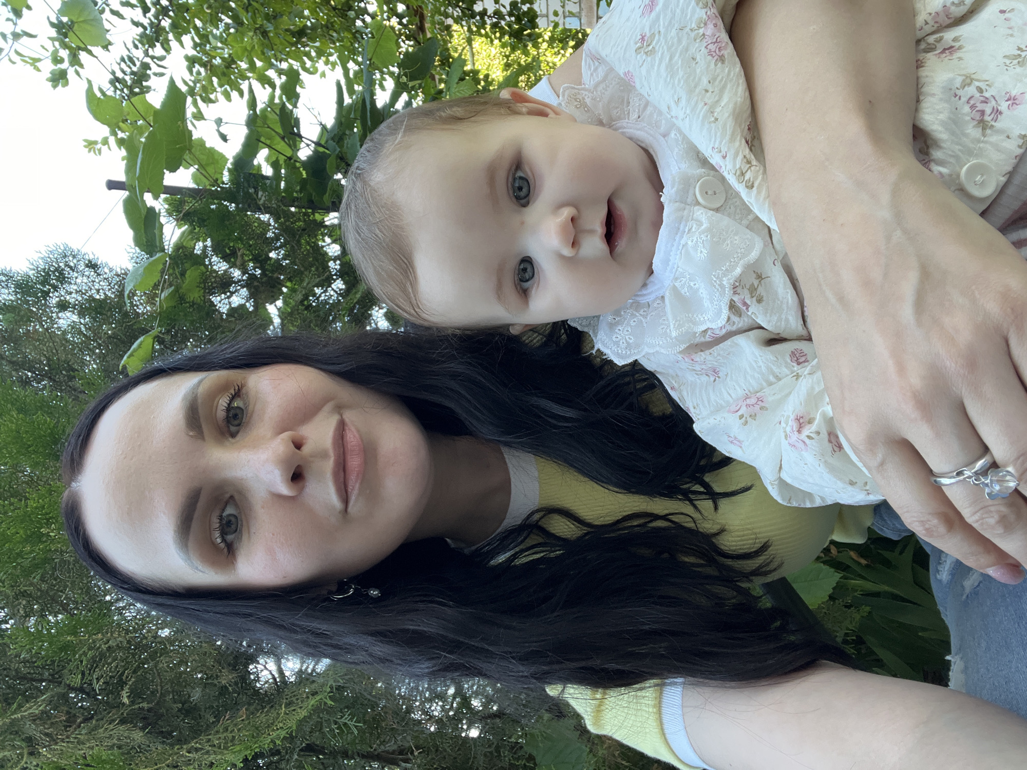 A woman and a baby are posing for a selfie outdoors. The woman, with long dark hair and wearing a yellow shirt with a white collar, is holding the baby, who is dressed in a white floral outfit with lace trim. They are positioned in front of a backdrop of lush green foliage and trees, suggesting a natural, possibly park-like setting. The lighting indicates it is daytime. The woman has makeup on, and both subjects appear to be looking towards the camera.