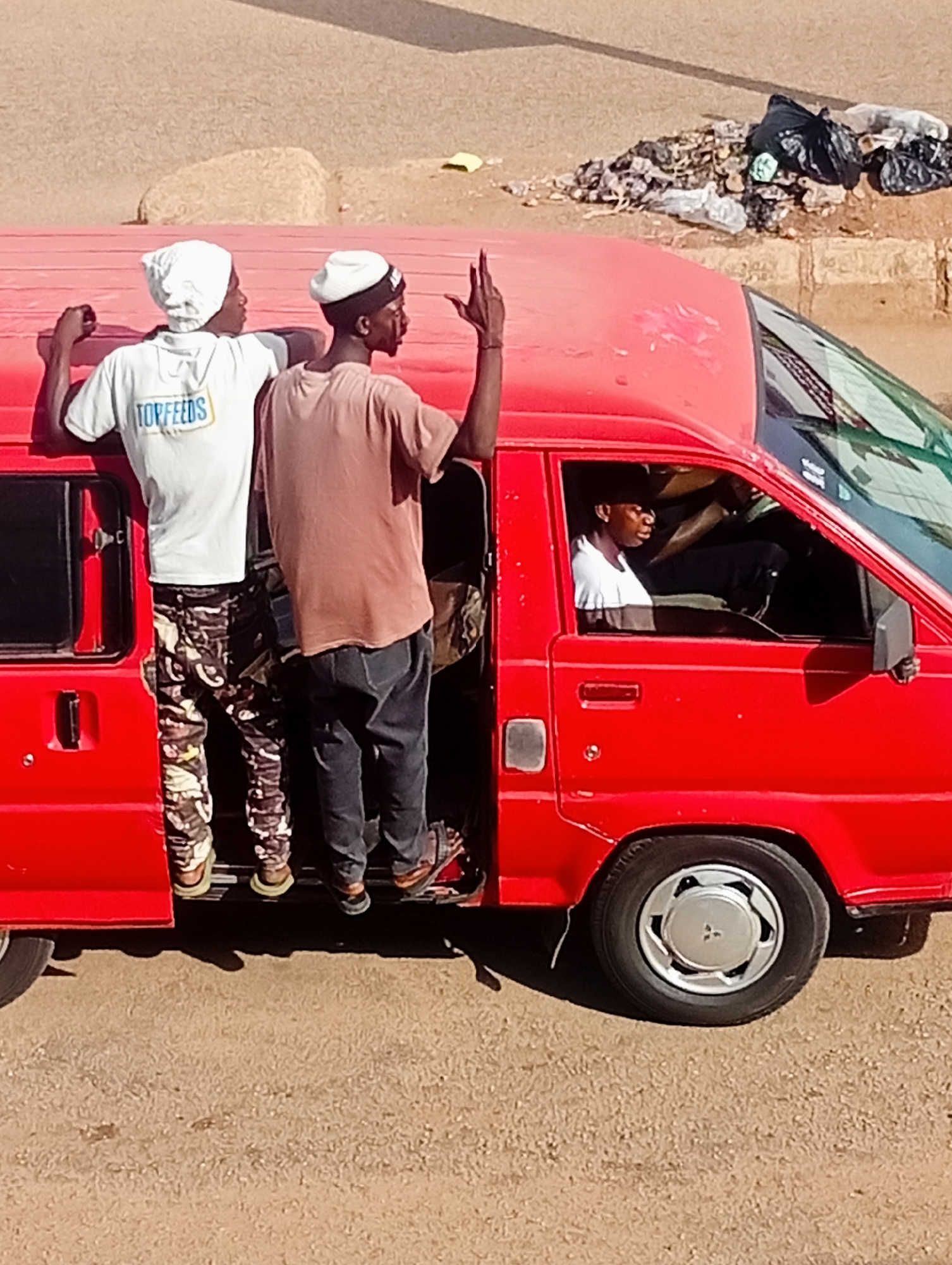 In Bukuru, Nigeria, a red van is parked on a dirt road next to a pile of trash. Two young men are standing in the open doorway of the van, with one reaching a hand out. A third young man is visible inside the van, in the driver's seat. The scene appears to be in a somewhat disheveled urban or semi-urban environment.