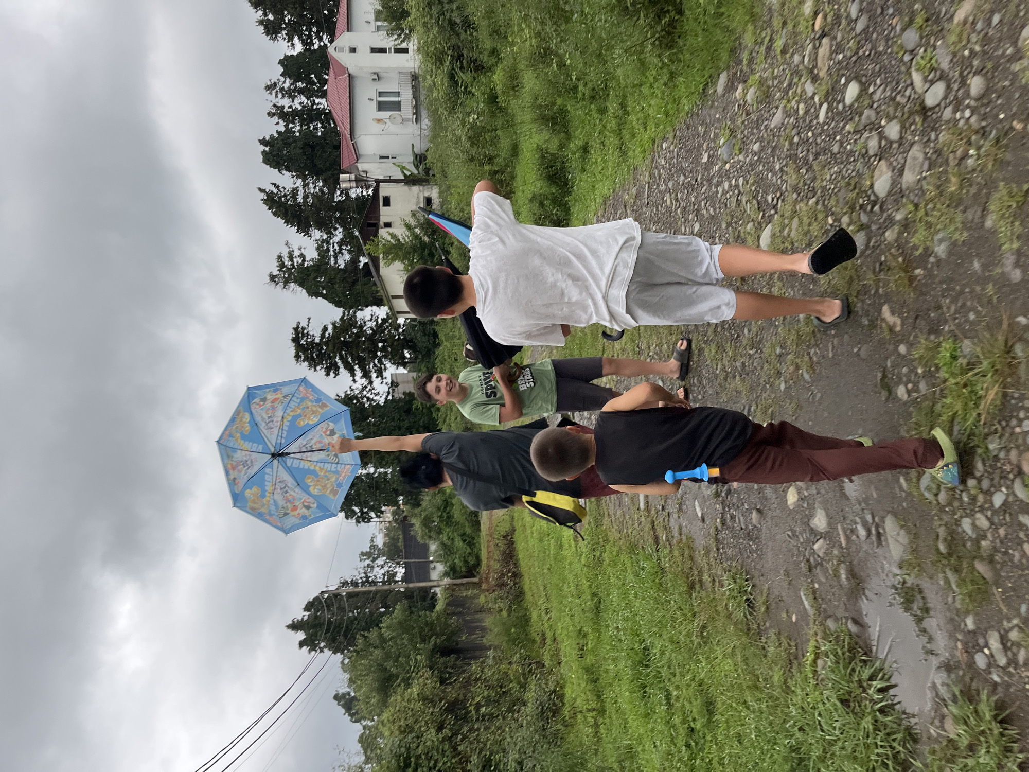 Four individuals are walking on a gravel path in Ureki, Georgia. A person in the back is holding a blue umbrella with cartoon characters on it. The person in the front is wearing a black tank top and brown pants with a blue water gun tucked into their waistband. The middle person is wearing a green t-shirt and dark shorts. The person at the very front is wearing a white t-shirt and white shorts. The environment is overgrown with green grass and trees, and the sky is overcast. There are some puddles on the path, indicating recent rain. In the background, there are white houses with red roofs.