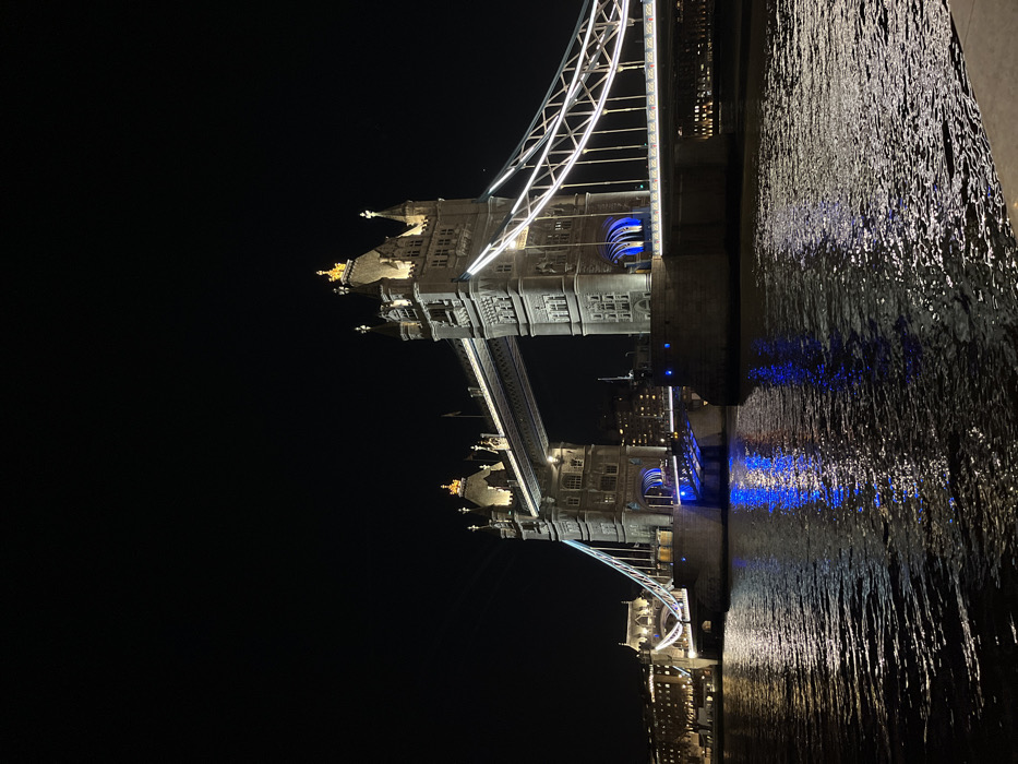 Tower Bridge in London is illuminated at night, reflected in the Thames River. The bridge's towers are detailed with golden embellishments and blue lighting. The sky behind the bridge is dark. The water's surface shimmers with the bridge's reflected lights, creating a dynamic texture. The image captures the iconic landmark's grandeur against the nighttime backdrop.