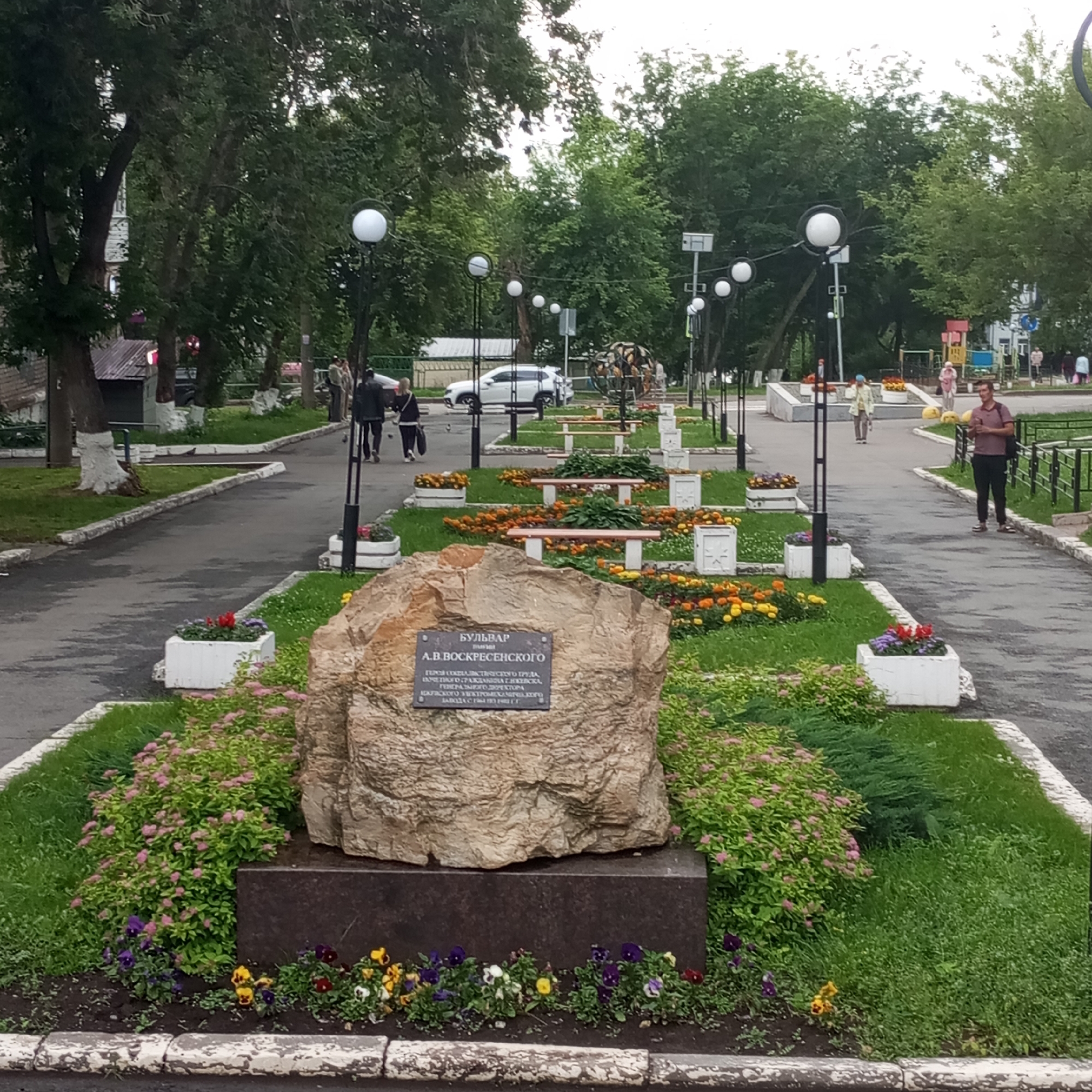 In Izhevsk, Russia, a memorial stone stands in a park-like setting, likely a boulevard named after A.V. Voskresensky. The stone, embedded with a dark plaque bearing Russian text, is surrounded by manicured flower beds featuring an array of colorful flowers, including pansies and vibrant pink and yellow blooms. The ground around the stone is covered in lush green grass, with a paved walkway curving through the scene. Several people are present, walking and standing, interacting subtly with the environment and each other. The background shows trees, streetlights with spherical globes, and what appears to be a playground, suggesting a public recreational area within the city.
