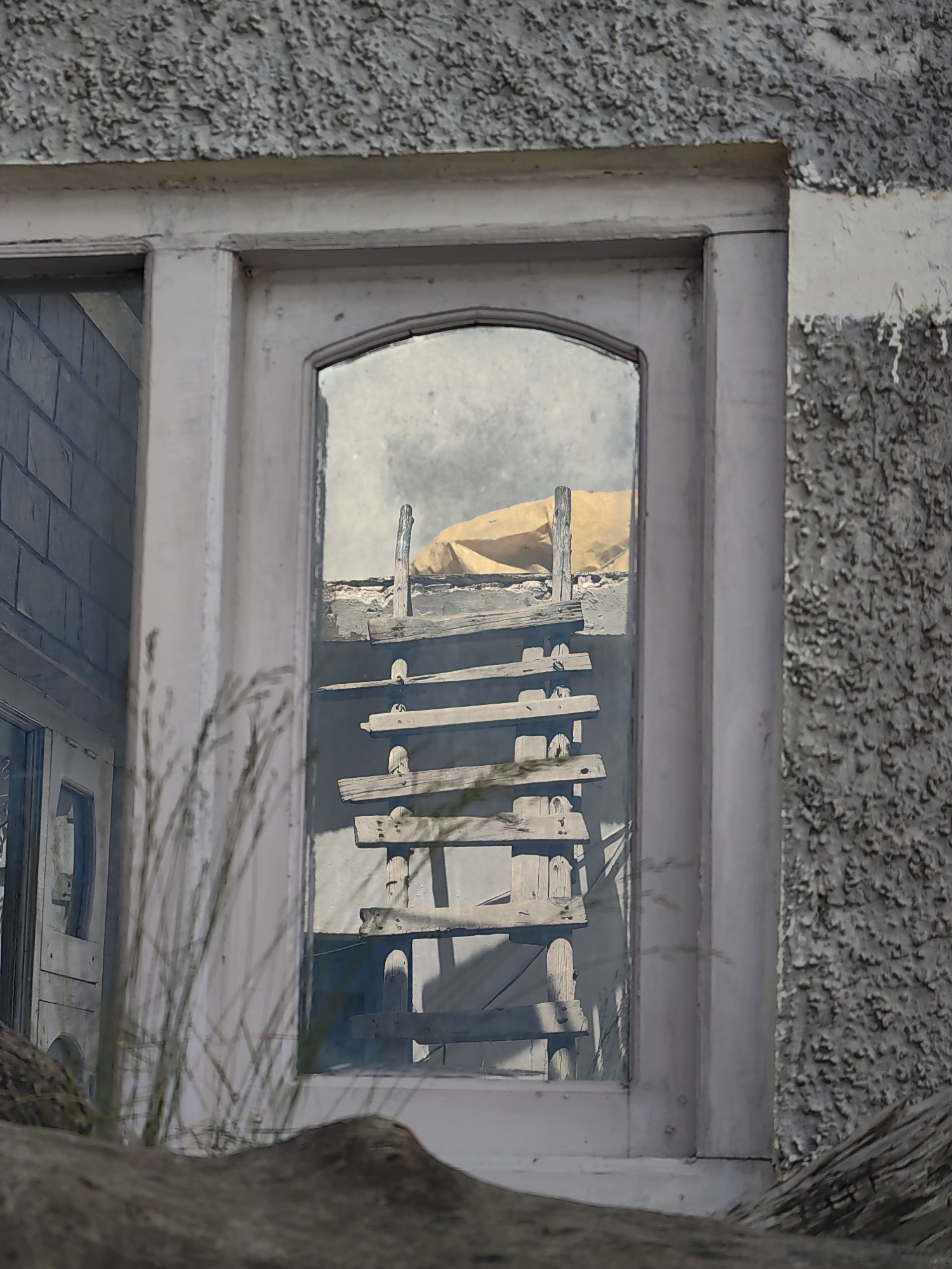 The image captures a close-up view of a window in Kishorl, India. The window frame is painted a light, muted color and appears to be made of wood, showing signs of wear and peeling paint. The window itself is a glass pane, reflecting the exterior environment.
Inside the window's reflection, a makeshift structure is visible. It seems to be constructed from rough-hewn wooden planks, forming a tiered or ladder-like arrangement. These planks are weathered and have exposed nails. Above the wooden structure, a large piece of tan-colored material, possibly a tarp or fabric, is draped and secured by a vertical wooden post. The background in the reflection is hazy and appears to be an outdoor, possibly rural or industrial, setting with a textured grey surface.
To the left of the main window, another section of the building is visible, featuring a door and a wall constructed with what looks like blue-grey brickwork. The foreground of the image is occupied by blurry, dry grass stalks and the rough texture of a stone or concrete structure. The overall impression is one of an old, possibly abandoned or disused building with visible signs of decay and improvisation in its construction.