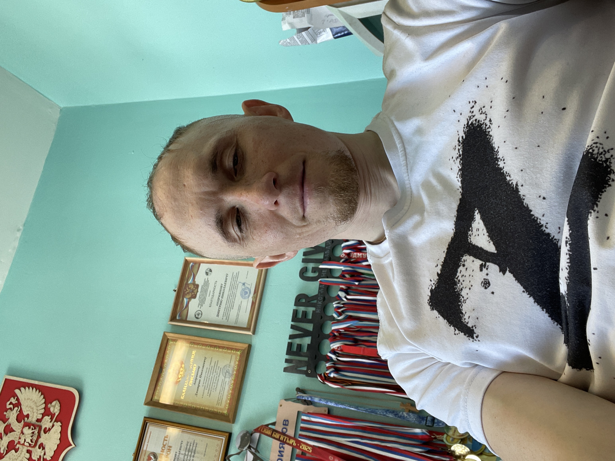A selfie captures a man indoors, positioned against a turquoise wall adorned with framed certificates, the Russian coat of arms, a medal holder with the inscription "NEVER GIVE", and hanging medals. The certificates feature Cyrillic script. The medal holder also has Cyrillic text indicating "UDMU". A tag reading "СКИЙ БОГАТЫРЬ - 2025" is visible on a red, white, and blue medal ribbon. The man is wearing a white t-shirt with a large black design. The location is likely Sarapul, Russia, indicated by "UDMU" and the Russian coat of arms.
