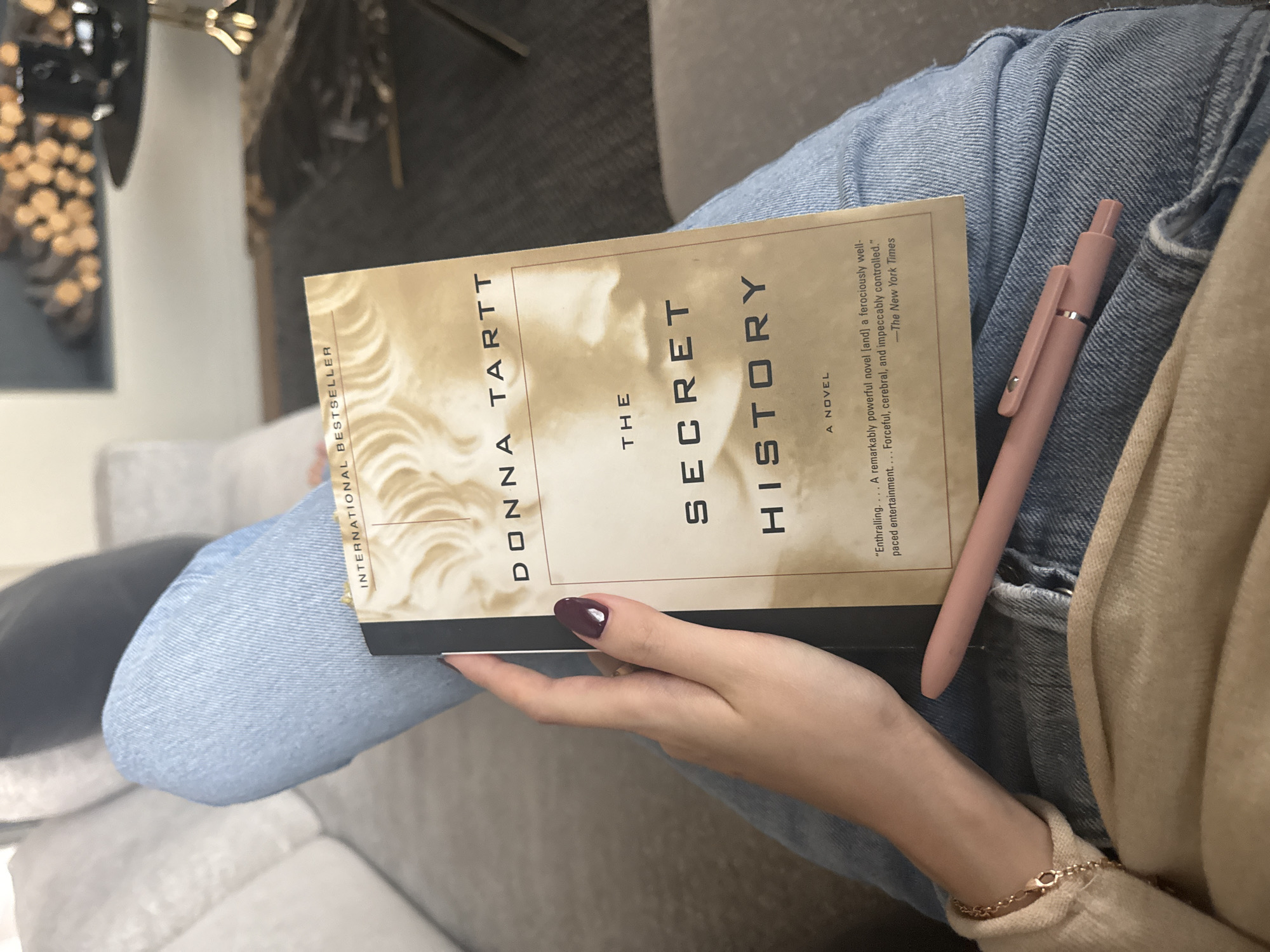 A person wearing light blue jeans and a beige sweater is seated on a light gray couch and is holding the book "The Secret History" by Donna Tartt. A pink pen rests on the jeans next to the book. The book is positioned at an angle so the title and author are visible. The person's hand is adorned with a dark nail polish and a gold bracelet. The photo was taken indoors.