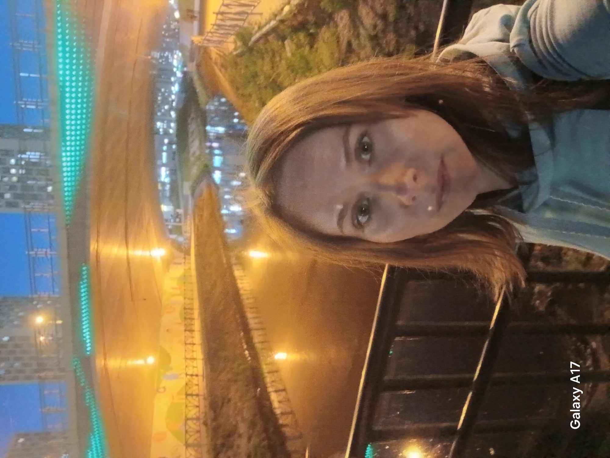 A woman is standing on a bridge at night, taking a selfie. The location is Yekaterinburg, Russia, as indicated by background elements. The bridge has black metal railings. Behind the railing is a highway/roadway with rows of green LED lights along the median and yellow street lights lining the sides. On the left of the road are tall buildings, illuminated with lights. On the right side of the roadway, there's a mural or design element. The woman is wearing a light blue top with a hood. The phone used to take the picture is a Galaxy A17, as indicated in the bottom right corner of the frame.