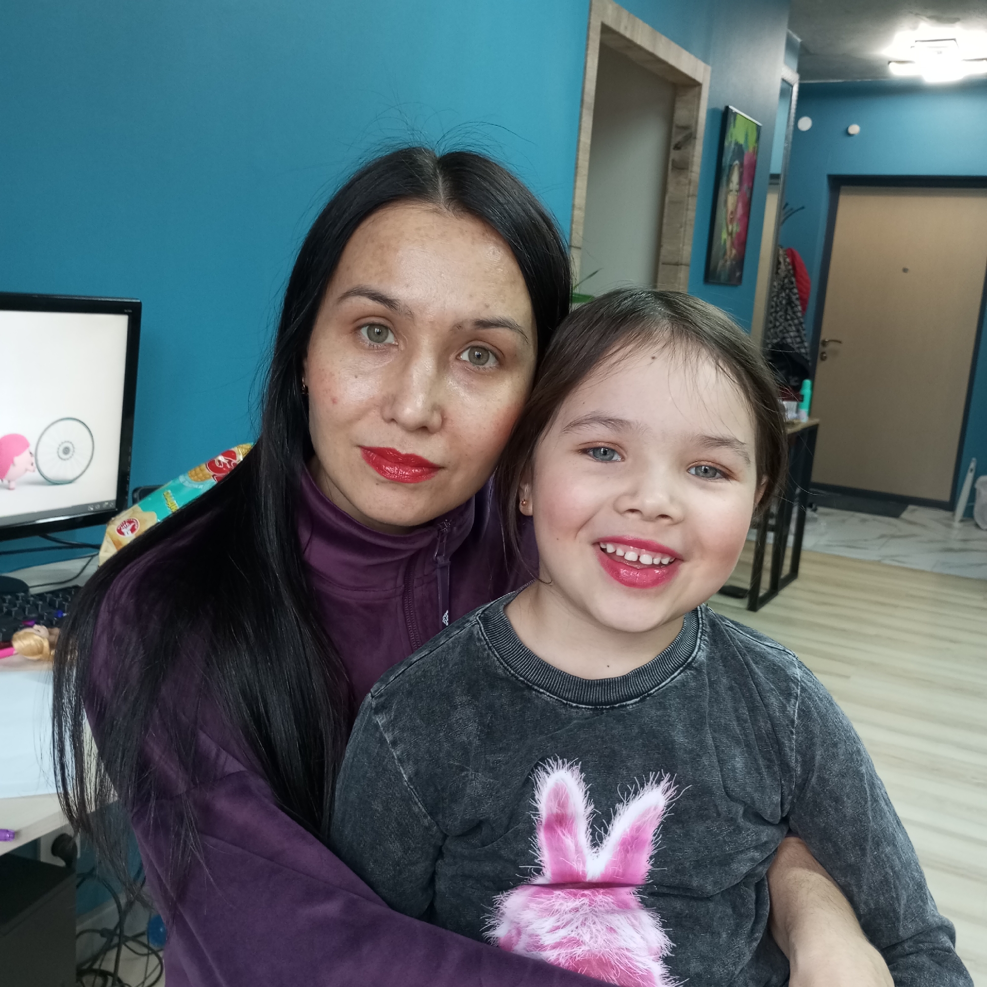 In an indoor setting, a woman and a child pose for a photo. The woman, with long dark hair and red lipstick, wears a purple top. The child, with light eyes and red lipstick, is wearing a dark grey shirt with a pink bunny graphic. A computer monitor is visible in the background. The location is likely inside a home in Pervomaysky, Russia, based on the request, although there are no specific details within the image to confirm this.