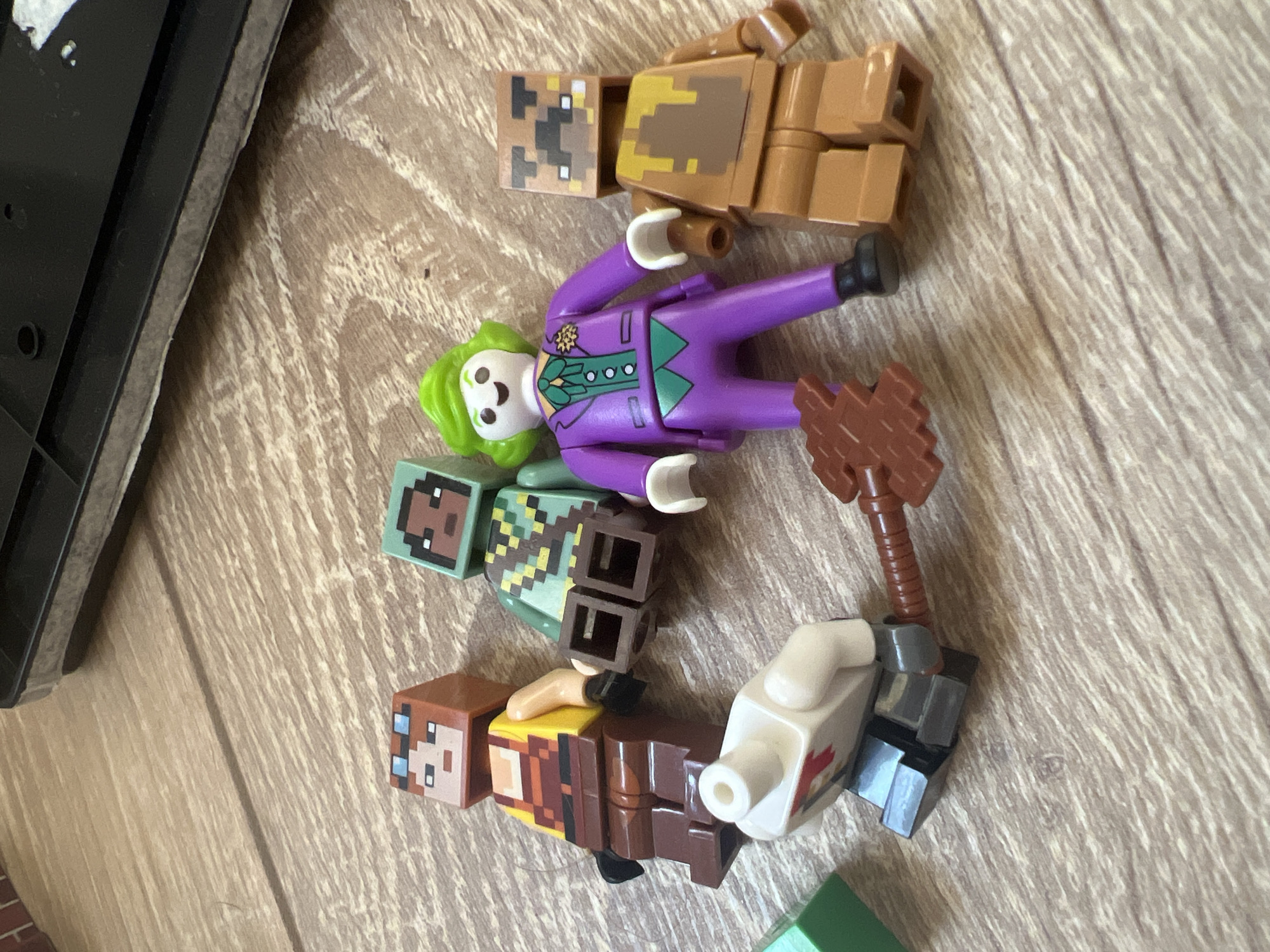 Six figures, possibly toys, are arranged on a wooden surface. From left to right: a tan-skinned figure with orange hair and blue goggles; a green-skinned figure with a patterned torso; a figure dressed as the Joker in a purple suit; a brown figure with a pixelated face and a dripping brown substance on its torso; a white figure with a red symbol on its chest; and a brown weapon with a textured handle. The figures are not in any discernible city environment.