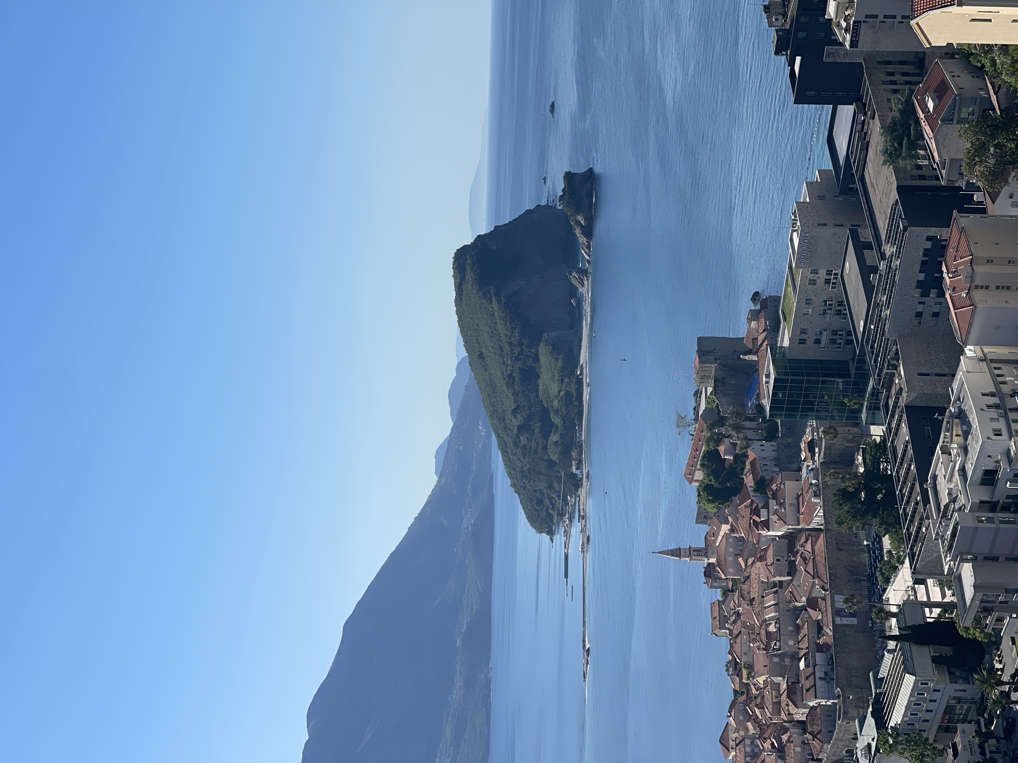 The image shows an elevated view of Budva, Montenegro, including the old town and coastal scenery. The old town is characterized by buildings with red-tiled roofs, including a church steeple visible amongst the tightly packed structures. To the right of the old town, modern buildings and hotels are observed. The blue Adriatic Sea stretches out to the horizon, and Sveti Stefan islet is also seen. A large mountain range sits behind the coastline, and the sky is clear and blue.