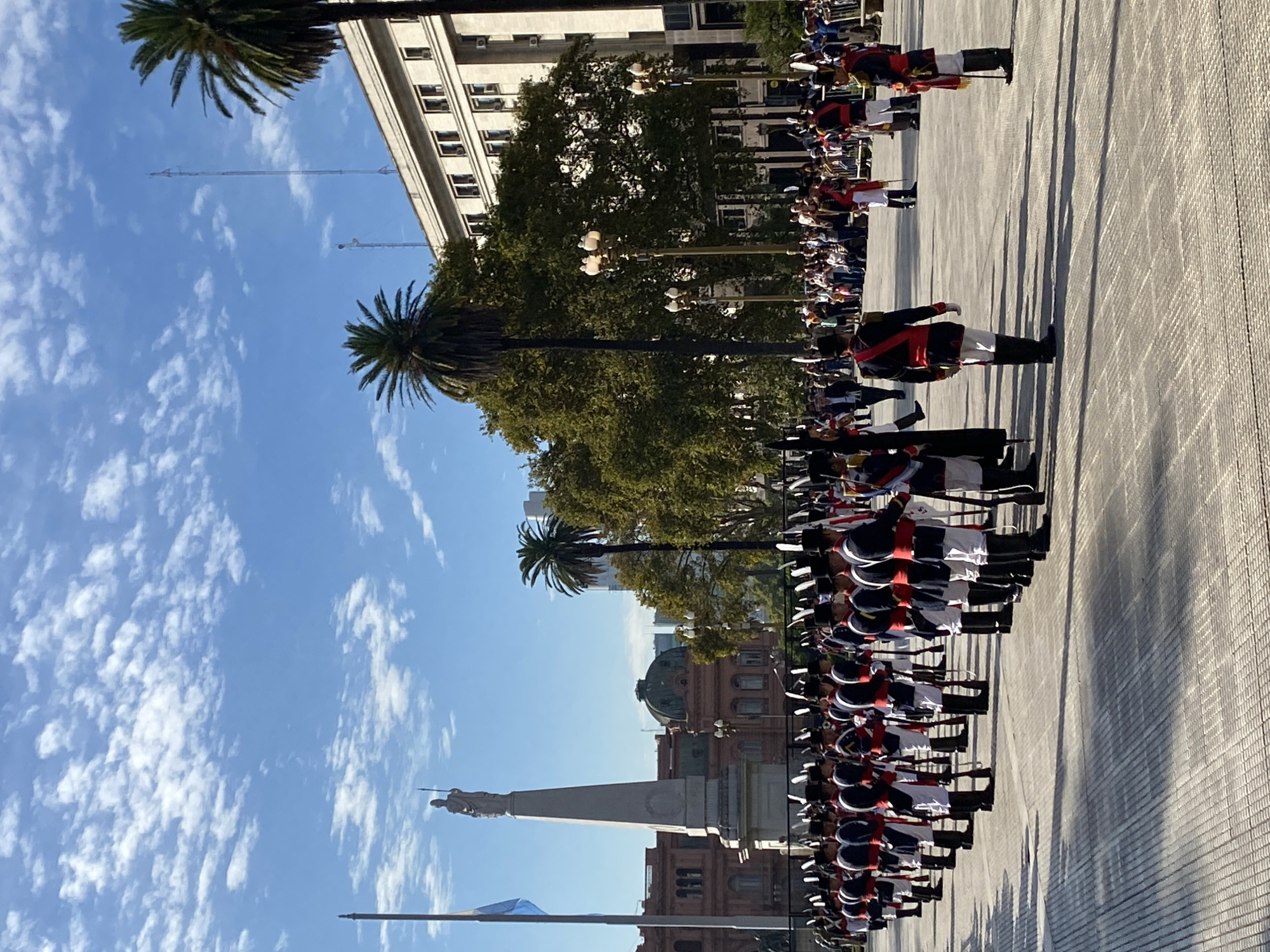 A medium, horizontally-oriented photograph shows a line of soldiers standing at attention in the Plaza de Mayo, Buenos Aires, Argentina. The soldiers are uniformly dressed in black boots and pants, white shirts, red belts and epaulettes, and black hats with white plumes. They are holding rifles. In the background, the Pirámide de Mayo monument and the Casa Rosada government building are visible. Palm trees are also visible, as is a building and the blue sky with wispy clouds. The paved ground is gray and beige.