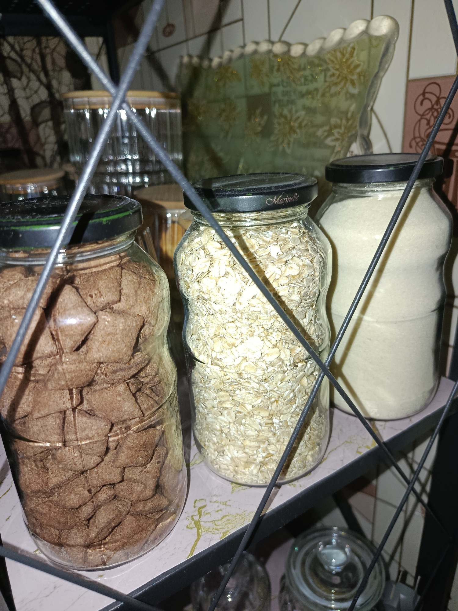 The image displays three glass jars filled with food items on a shelf, partially obscured by black wire shelving. The leftmost jar contains brown, square-shaped food items, possibly cereal or crackers. The middle jar holds rolled oats. The rightmost jar contains a white granular substance, likely flour or sugar. All jars have black lids. In the background, out of focus, are other kitchen items, including decorative jars and a patterned wall or backsplash. The overall setting appears to be a kitchen or pantry. There is no information in the image to suggest a specific location in Tashkent, Uzbekistan.
