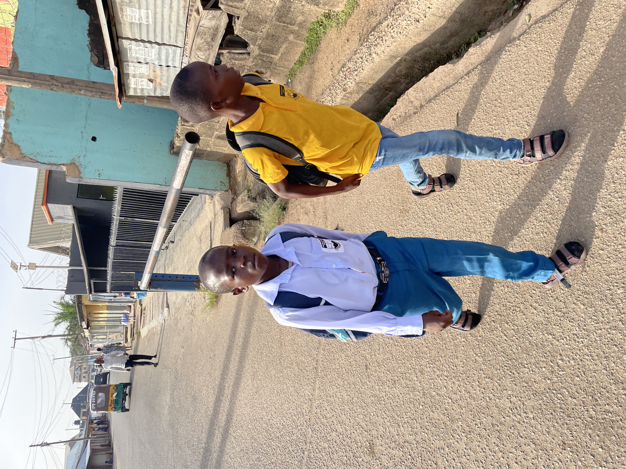 The image shows two young boys standing on a paved road, likely in an urban or semi-urban setting, under bright sunlight, indicated by strong shadows. The image appears to be rotated 90 degrees clockwise.
The boy in the foreground wears a white long-sleeved shirt with a visible school emblem on the left chest pocket, blue or teal trousers, and brown sandals. He carries a dark blue backpack and looks directly at the viewer with a neutral expression.
Behind him, slightly to his right, stands another boy wearing a yellow polo shirt with a dark logo on the left chest, blue jeans, and dark brown sandals. He also carries a dark backpack and looks towards the left side of the frame. Both boys have short, dark hair.
The setting includes buildings with painted walls, some featuring corrugated iron roofs, visible on the left and behind the boys. A turquoise-painted wall is prominent in the mid-ground. Utility poles with numerous wires run across the upper left portion of the image.
In the background, further down the road, an adult male figure is walking, and a green and yellow auto-rickshaw (commonly known as a "keke napep" in some regions) is visible. The license plate or identification on the auto-rickshaw seems to have numbers and letters, possibly "PT 18245", but it's not fully legible. The road surface is granular and light-colored. The overall impression is that of a bustling street scene in a sunny, possibly West African, environment, with the boys either heading to or returning from school. No other clear text is legible.