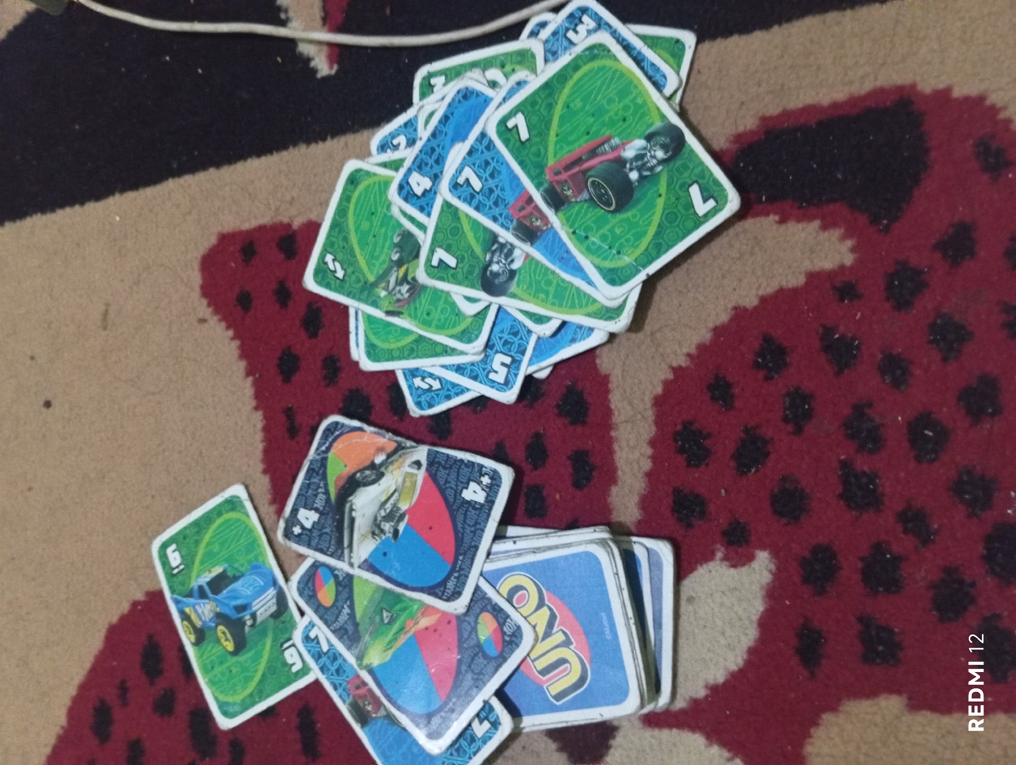 In Pagar Alam, Indonesia, a collection of Uno cards, specifically from the Hot Wheels edition, are scattered on a patterned rug. The rug features a dominant red base with black and tan organic shapes resembling animal spots or abstract floral elements. The Uno cards are of various colors and designs, with several depicting race cars. Some cards are face up, revealing numbers and symbols such as '4', '7', and '5', along with the distinctive Uno logo on a blue card. Other cards are partially obscured or face down. The lighting suggests an indoor setting. The bottom left corner of the image displays the text "REDMI 12".