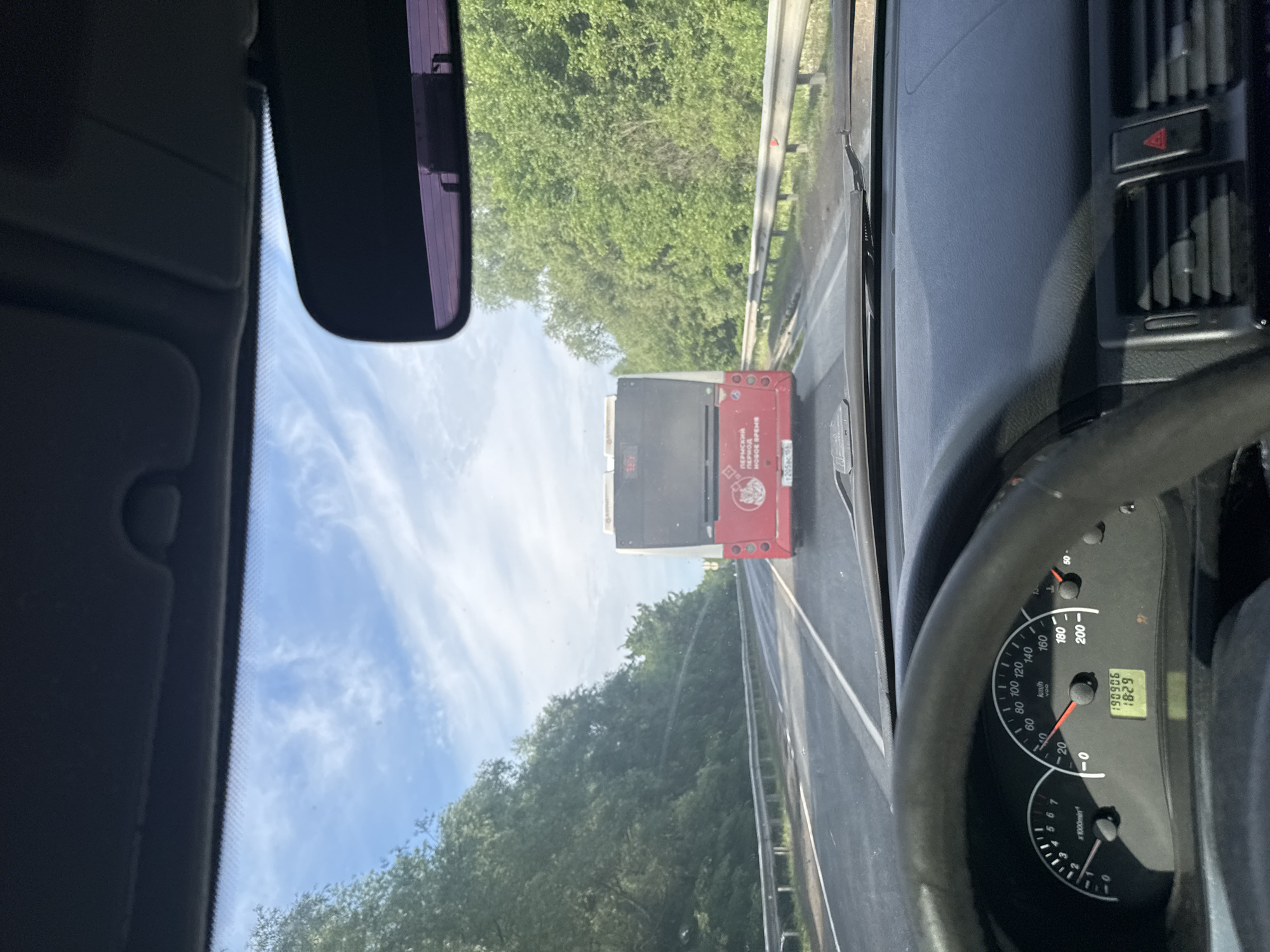 The image presents a first-person view from inside a moving vehicle, looking forward through the windshield. The scene depicts a road in Усть-Сыны, Russia, on a bright, sunny day with a blue sky and scattered white clouds.
In the foreground, elements of the car's interior are visible. On the right, a portion of the steering wheel and the dashboard instrumentation are seen, including a speedometer showing a speed of approximately 40-50 km/h and an odometer reading 198906 km. A digital display indicates "1829", likely a trip meter. A red triangular hazard light button is also visible on the dashboard. On the left, the car's rearview mirror hangs, reflecting a subtle purple tint.
The primary subject outside the car is a red bus ahead, which the car is following. Its rear is clearly visible. The roadside is lined with dense green trees and a metal guardrail.
Visible text on the bus includes "ПЕРМСКИЙ", "ПЕРИОД", and "НОВОЕ ВРЕМЯ", which translates to "Permian Period New Time" or "Perm Region, New Time," indicating the bus's operational area is likely the Perm region of Russia. A vehicle number or license plate "1205ac1" is also present on the bus. The car's dashboard features standard numerical markings for speed (0-200 km/h) and engine RPM (x1000min).