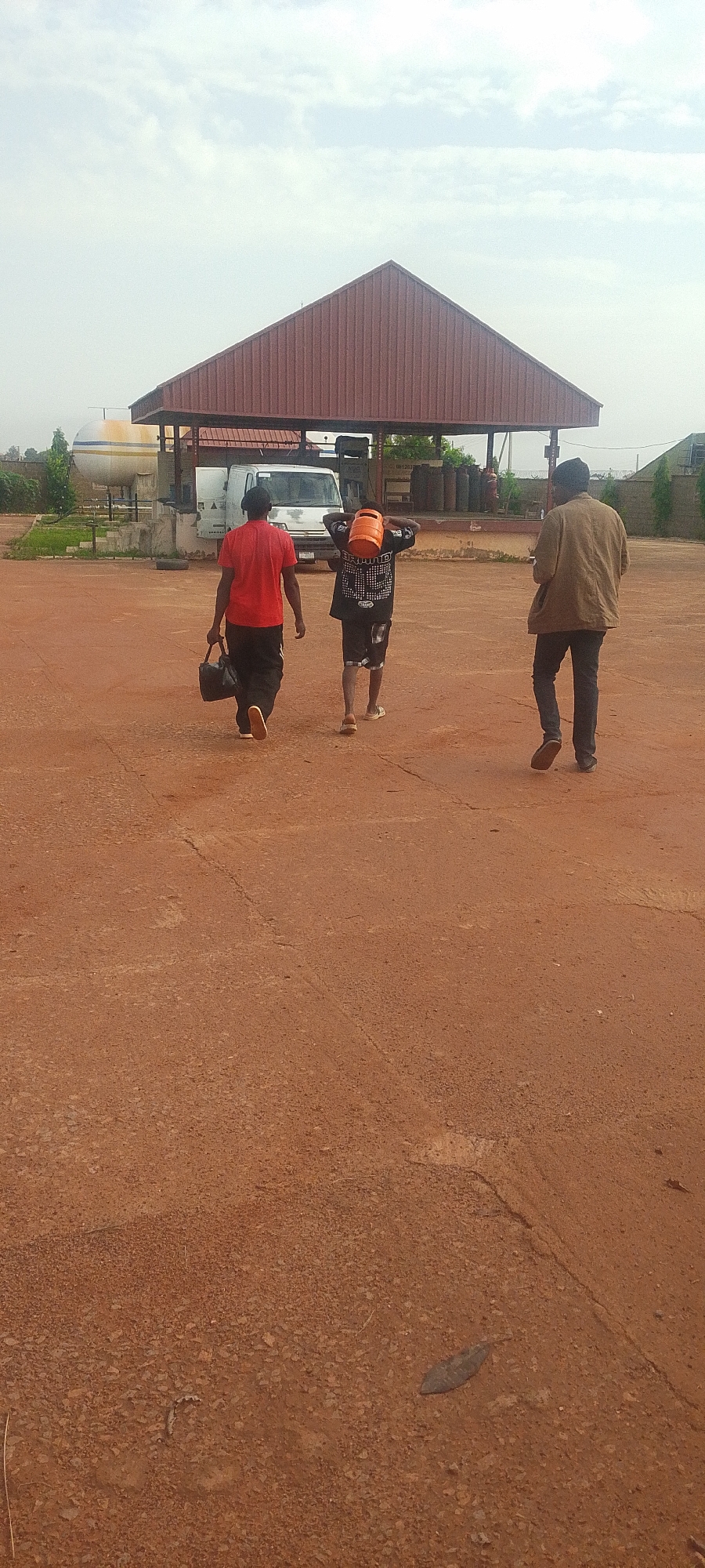 Three individuals are walking away from the camera in an outdoor, open area with a reddish-brown, gravelly surface. In the background, a large, red, gabled structure stands, providing cover over a white truck and other equipment. To the left of the structure, a large, white cylindrical tank is visible. The sky above is cloudy.
The person on the left is wearing a red t-shirt and black pants, carrying a black bag in their left hand. The person in the middle is wearing a black t-shirt with white lettering and black shorts, carrying an orange propane tank on their shoulders. The person on the right is wearing a brown jacket and dark pants.
The scene appears to be a commercial or industrial area, possibly a gas station or depot, given the presence of the propane tank, truck, and cylindrical tank. The location of Jos, Nigeria is not explicitly indicated in the image itself, but this description focuses on the visual elements present.