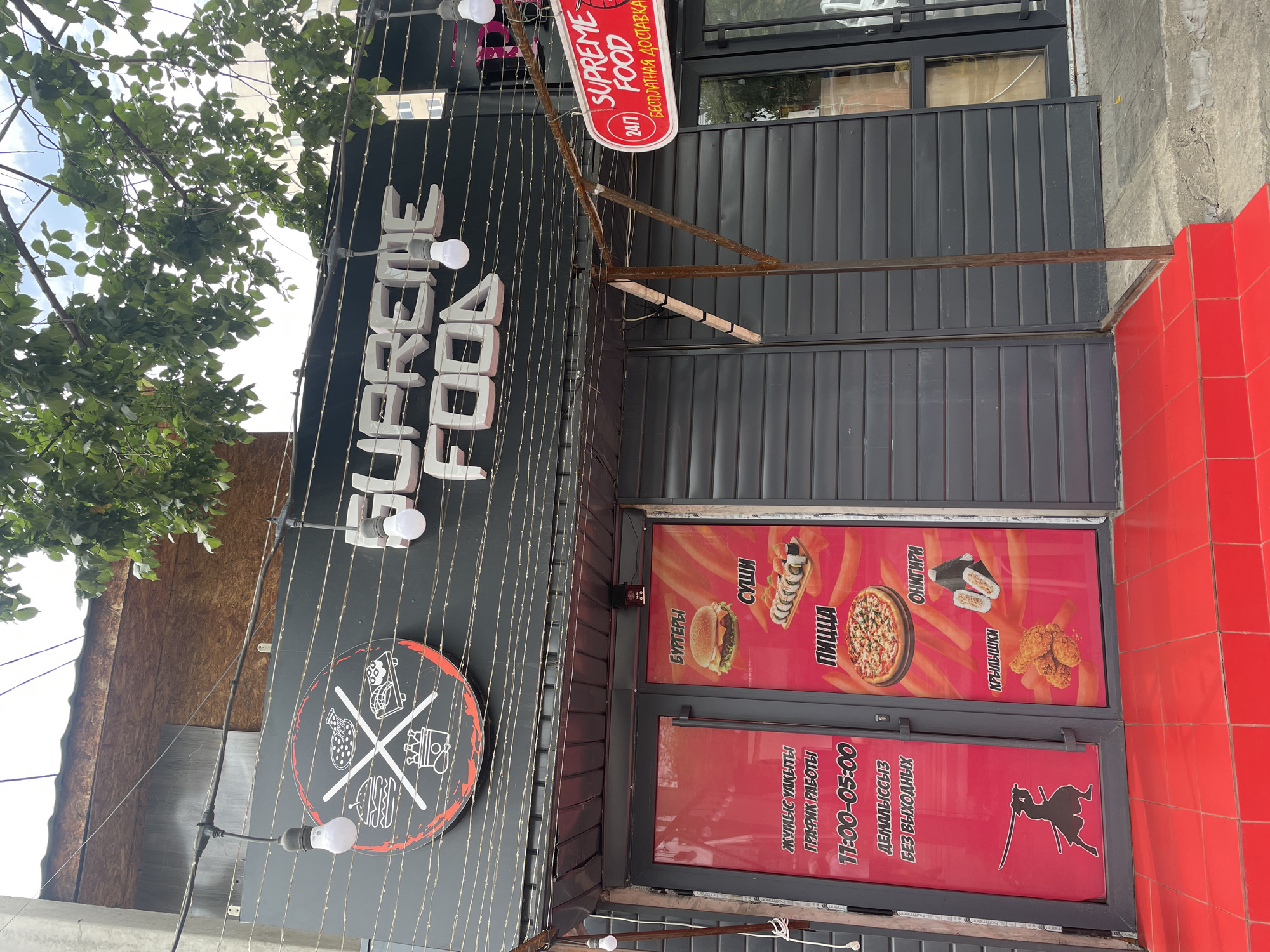 The image shows the exterior of a fast-food restaurant named "SUPREME FOOD" in Almaty, Kazakhstan. The restaurant has a dark gray facade with a prominent sign that reads "SUPREME FOOD" in large, white, 3D letters. The entrance is a double door with red panels featuring images of burgers, sushi, and pizza, along with text in Cyrillic script. To the right of the entrance, a red oval sign with "SUPREME FOOD" and "БЕСПЛАТНАЯ ДОСТАВКА" (free delivery) is visible. The restaurant is decorated with string lights and has a circular logo with icons representing food items. In front of the entrance, there are red tiled steps. The surrounding environment includes a tree with green leaves providing shade, a cloudy sky, and parts of other buildings in the background. There are no people visible in the image.