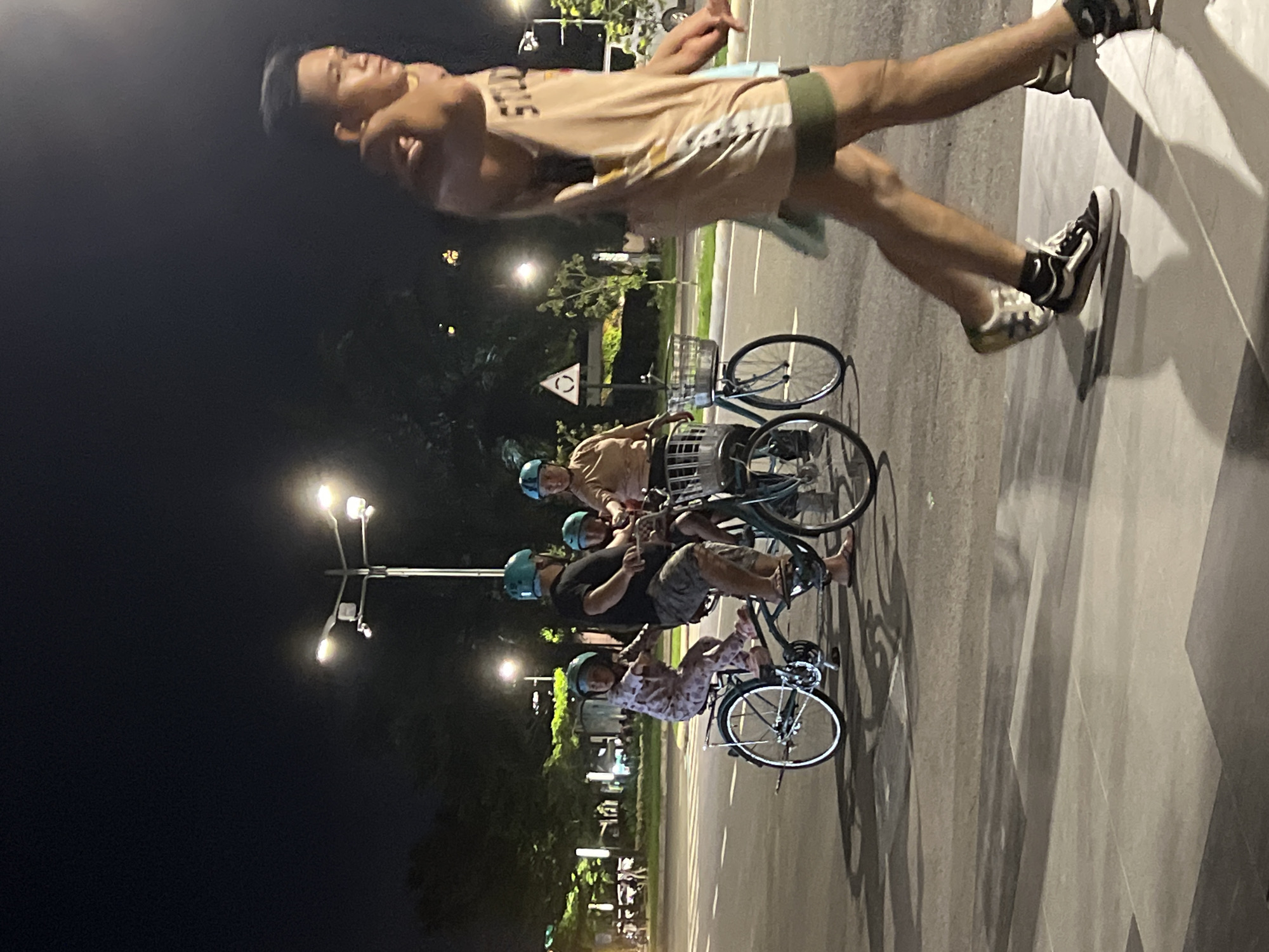 The image captures a nighttime scene in what appears to be Davao City, Philippines, showing a group of people engaged in a leisure activity with bicycles.
In the foreground, a man stands upright on a paved surface, his body angled sideways. He wears a light-colored sleeveless shirt with "DAVAO" partially visible on it, shorts with a green waistband, and sneakers. He appears to be balancing or posing, possibly for a photo.
Behind him, three other individuals are visible with bicycles. They are all wearing helmets, suggesting a focus on safety. One individual is seated on a bicycle, while another stands next to their bike. The bicycles feature front baskets. The group seems to be taking a break or riding together on a paved path or sidewalk adjacent to a road.
The setting is an outdoor area illuminated by several bright streetlights, casting shadows on the ground. Trees and foliage line the background, indicating a park, promenade, or well-landscaped urban area. A triangular road sign is visible in the middle ground. The time of day is clearly night due to the dark sky and reliance on artificial lighting. The overall atmosphere suggests a casual evening outing or exercise.
