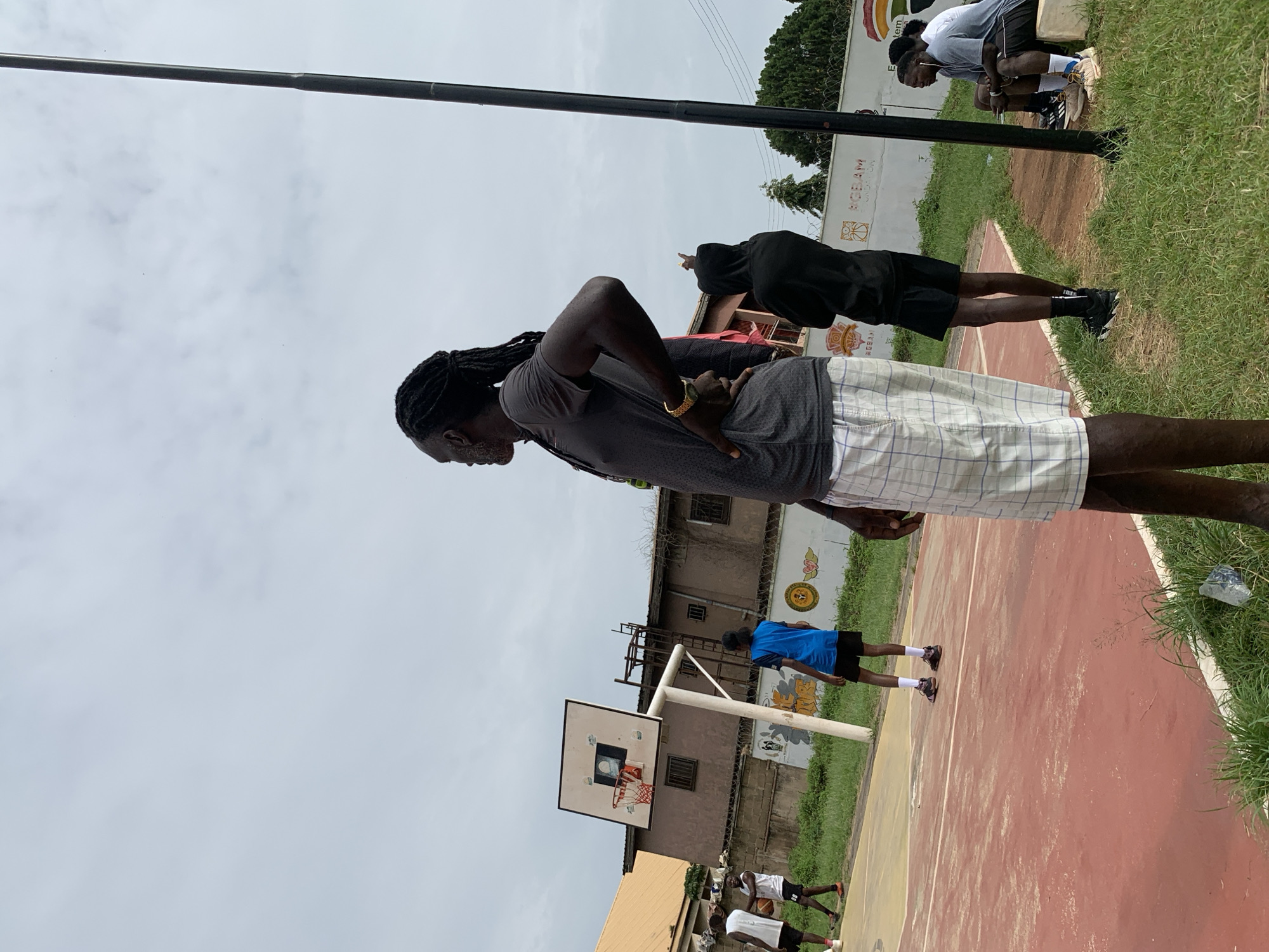 In Gura Top, Nigeria, an outdoor basketball court is visible under an overcast sky. The court features a red surface with white lines. Several individuals are present on and around the court. In the foreground, a tall man with dreadlocks, wearing a grey t-shirt and plaid shorts, stands with his hand on his hip, looking to the side. Behind him, another person in a black hooded jacket points upwards. On the court, two young men in athletic wear are playing basketball. One is wearing a white tank top and shorts, holding the ball, while the other, in a blue t-shirt and black shorts, stands nearby. Further in the background, a group of young men are seated on a bench, appearing to rest or watch the game. A basketball hoop with a white backboard and orange rim is positioned on the court. The surrounding environment includes green grass, trees, and buildings in the distance, suggesting an urban or semi-urban setting. A tall, dark pole, possibly a light post or part of a structure, stands prominently on the right side of the frame. The overall scene depicts a casual basketball game or practice session.