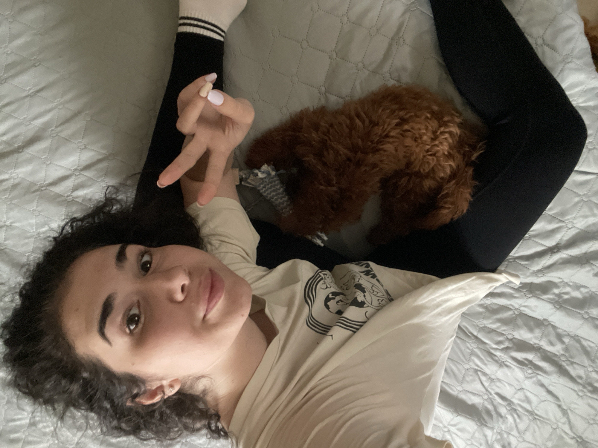 A young woman lies on a bed with a light gray, quilted bedspread. She is wearing black leggings and a white long-sleeved shirt with a black design on the front. Her dark curly hair is spread out on the bed. She is making a peace sign with her left hand. A small, brown dog is lying near her legs. The location is not evident from this image and cannot be determined to be Sofia, Bulgaria.