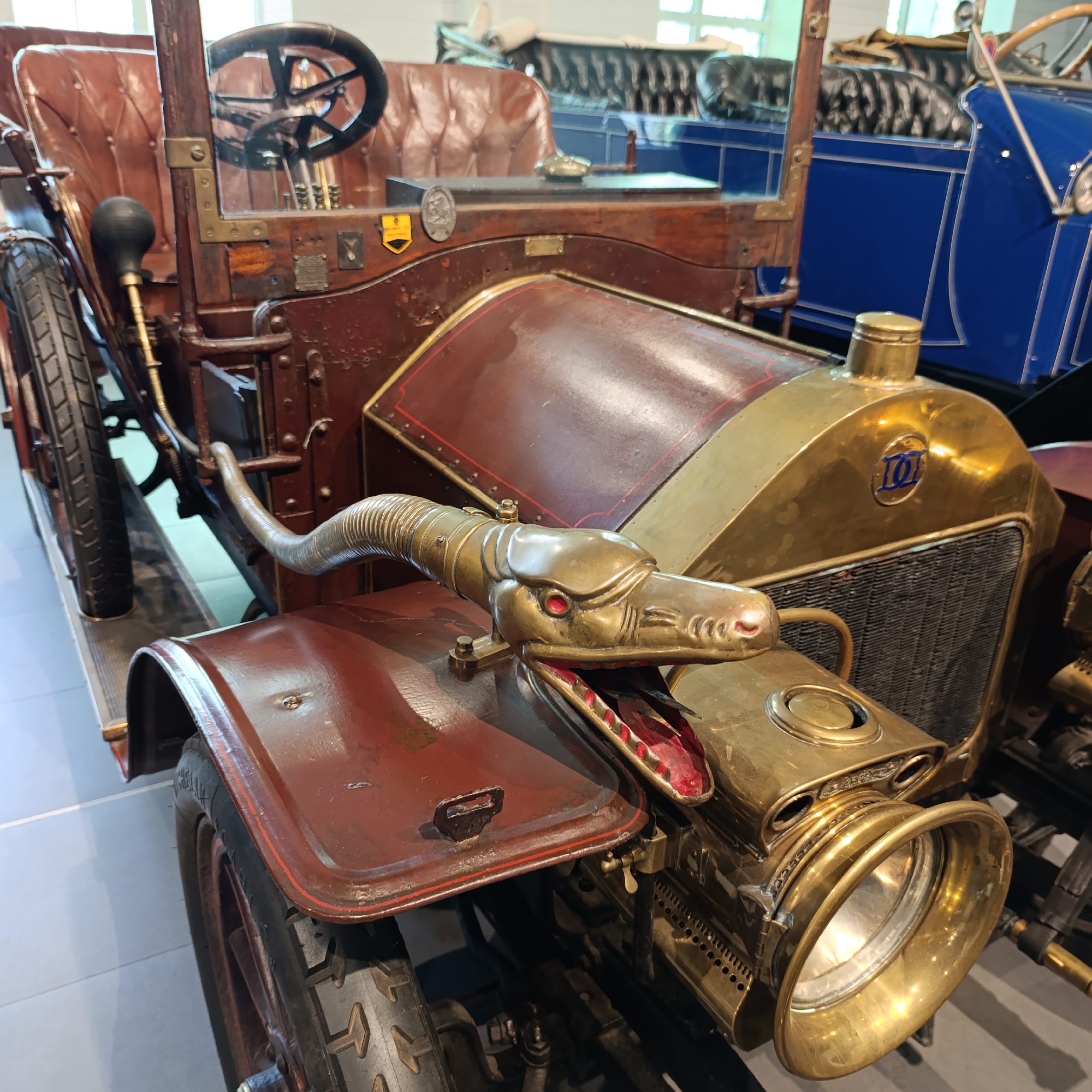 The image shows a vintage, possibly antique, car with unique features, including a dragon-shaped horn or ornament attached to the front near the headlight. The car has a brass-colored grill and headlight casing. The car's body is a combination of wood and possibly leather in brown and burgundy tones. It's located in Verkhnyaya Pyshma, Russia. The setting appears to be indoors, possibly a museum or exhibition hall. The environment is well-lit with other vehicles visible in the background.