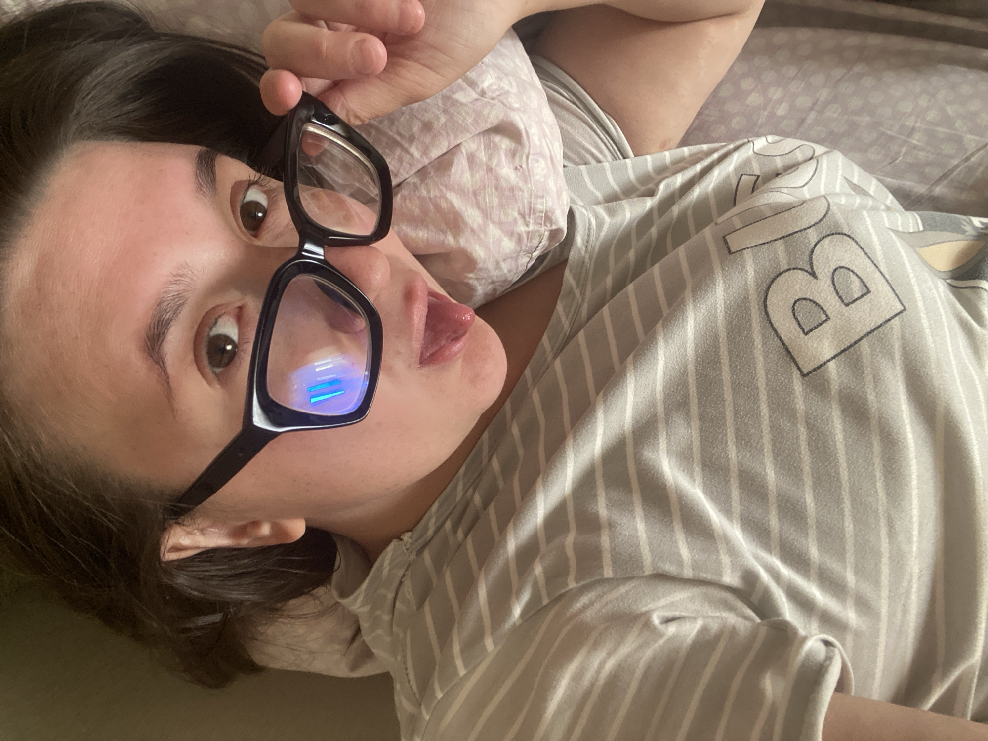 A close-up, high-angle shot captures a person with dark hair, lying down indoors, likely on a bed or couch. The individual is wearing a light grey and white striped shirt, which has partial letters printed on it, most notably a large "B". They are holding black-framed glasses up to their eyes with their right hand, looking directly at the camera. A playful expression is evident, with their tongue slightly extended. Bright blue and white rectangular reflections, possibly from a screen or artificial light source, are prominent on the lenses of the glasses. The background shows a light pink or patterned pillow and another grey sheet or blanket. The scene conveys a relaxed and informal moment, possibly a selfie taken during downtime. The location context provided is Chaikovsky, Russia.