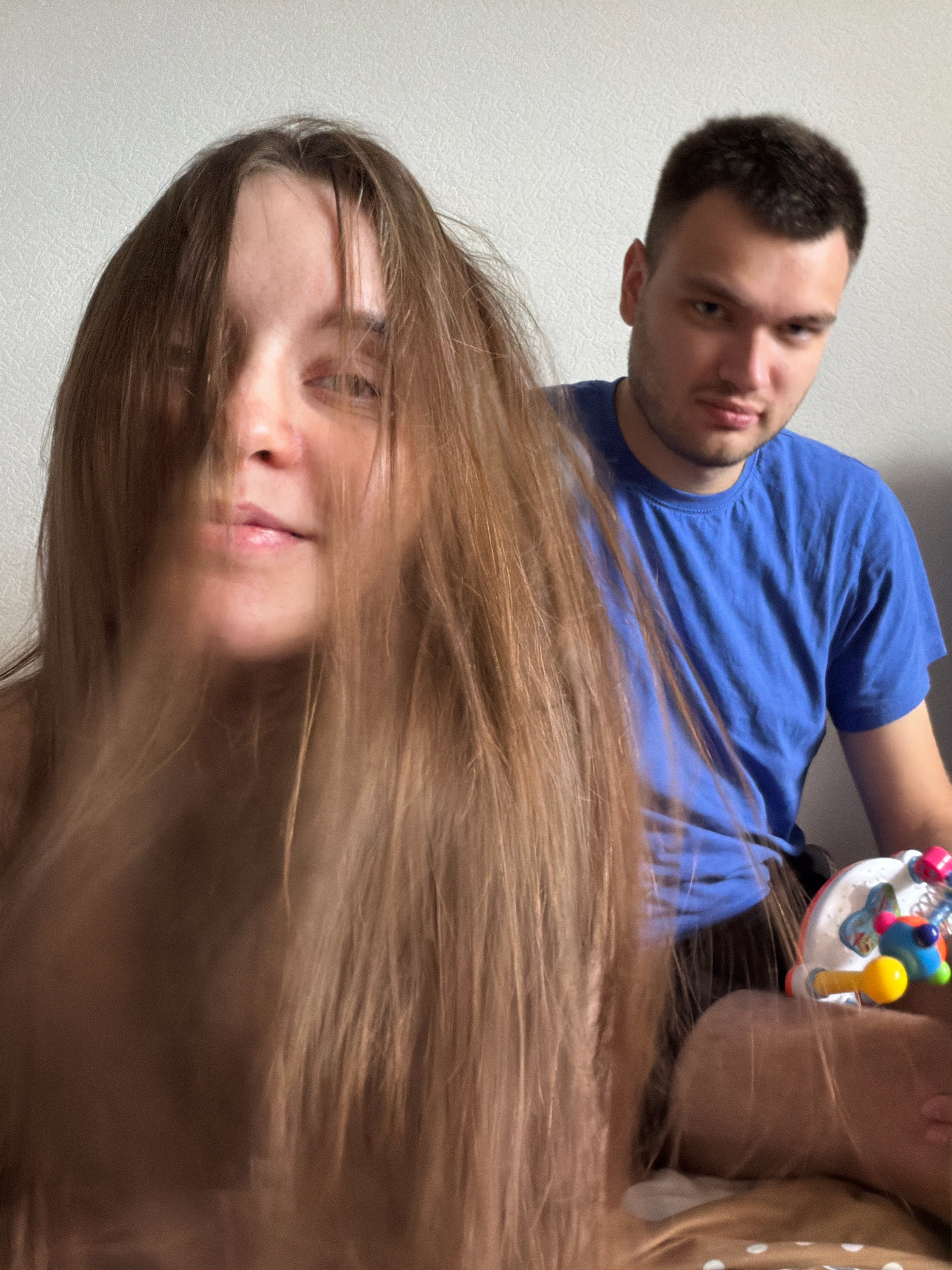 The image shows two people, a woman in the foreground and a man in the background. The woman's face is partially obscured by her long, light brown hair, but her eyes, nose, and mouth are visible. She is looking towards the camera. The man is to her right and slightly behind her. He has short brown hair and is wearing a blue t-shirt. He is looking at the camera with a neutral expression. In the lower right corner of the image, a colorful toy is visible. The background appears to be a wall and a bed. The lighting suggests an indoor setting. There is no information about the location in Noviy, Russia, associated with this image.