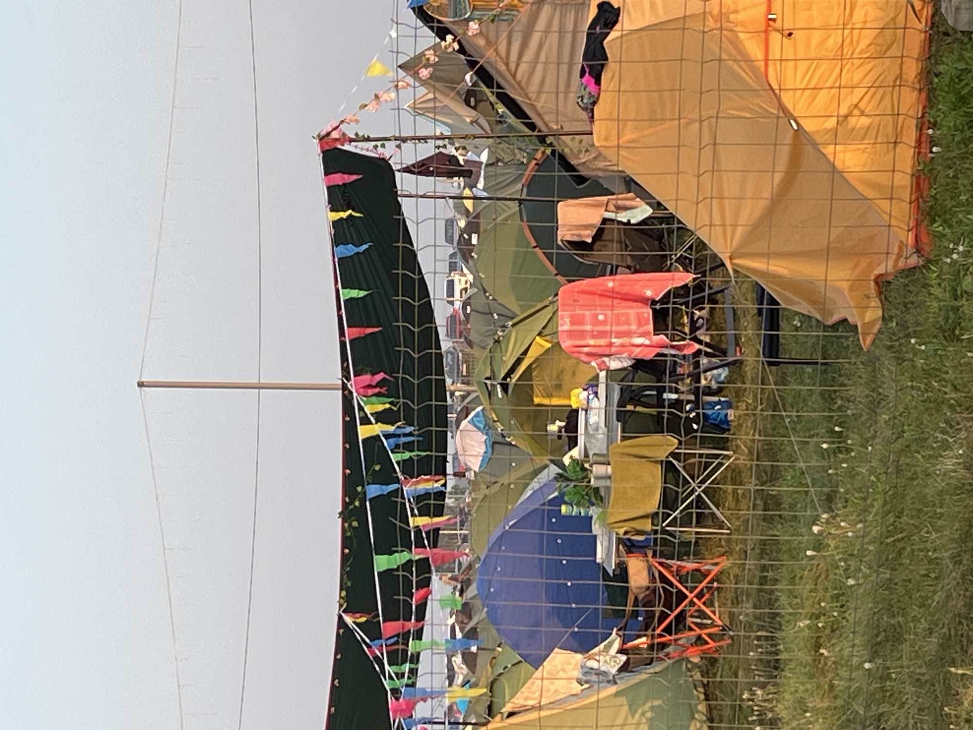 A camping site is depicted in Surakovo, Russia, under a clear sky. Numerous tents of various colors and sizes are pitched closely together, forming a dense encampment. A large green tarp is stretched overhead, adorned with colorful triangular flags and faux ivy, creating a festive atmosphere. A wire fence with a diamond pattern runs across the foreground, separating the viewer from the campsite. Inside the fenced area, a small table is set up with two folding chairs. A yellow tablecloth drapes over the table, and a plant sits on it, along with some containers. Various items of clothing, including a pink plaid cloth and a tan towel, are draped over the fence. In the background, more tents and a line of vehicles are visible. The ground is covered in grass, with scattered white wildflowers.