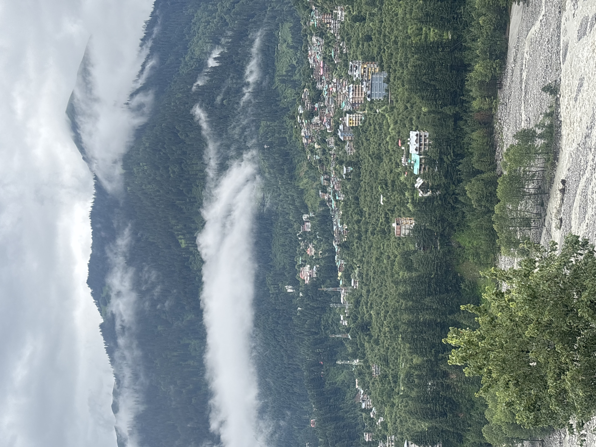 The image depicts a panoramic view of the town of Vashisht, India, nestled amidst lush green hills. In the foreground, a rocky riverbed with flowing water is visible, bordered by trees. The midground features a densely wooded hillside dotted with numerous buildings, indicating a residential area. These buildings exhibit a variety of architectural styles and colors, with some appearing to be hotels or guesthouses. Several prominent telecommunication towers stand among the buildings. The background is dominated by steep, mist-covered mountains covered in dense evergreen forests, with clouds partially obscuring the peaks, suggesting a mountainous, possibly monsoon-season, environment. The overall scene conveys a sense of a remote, picturesque mountain town.