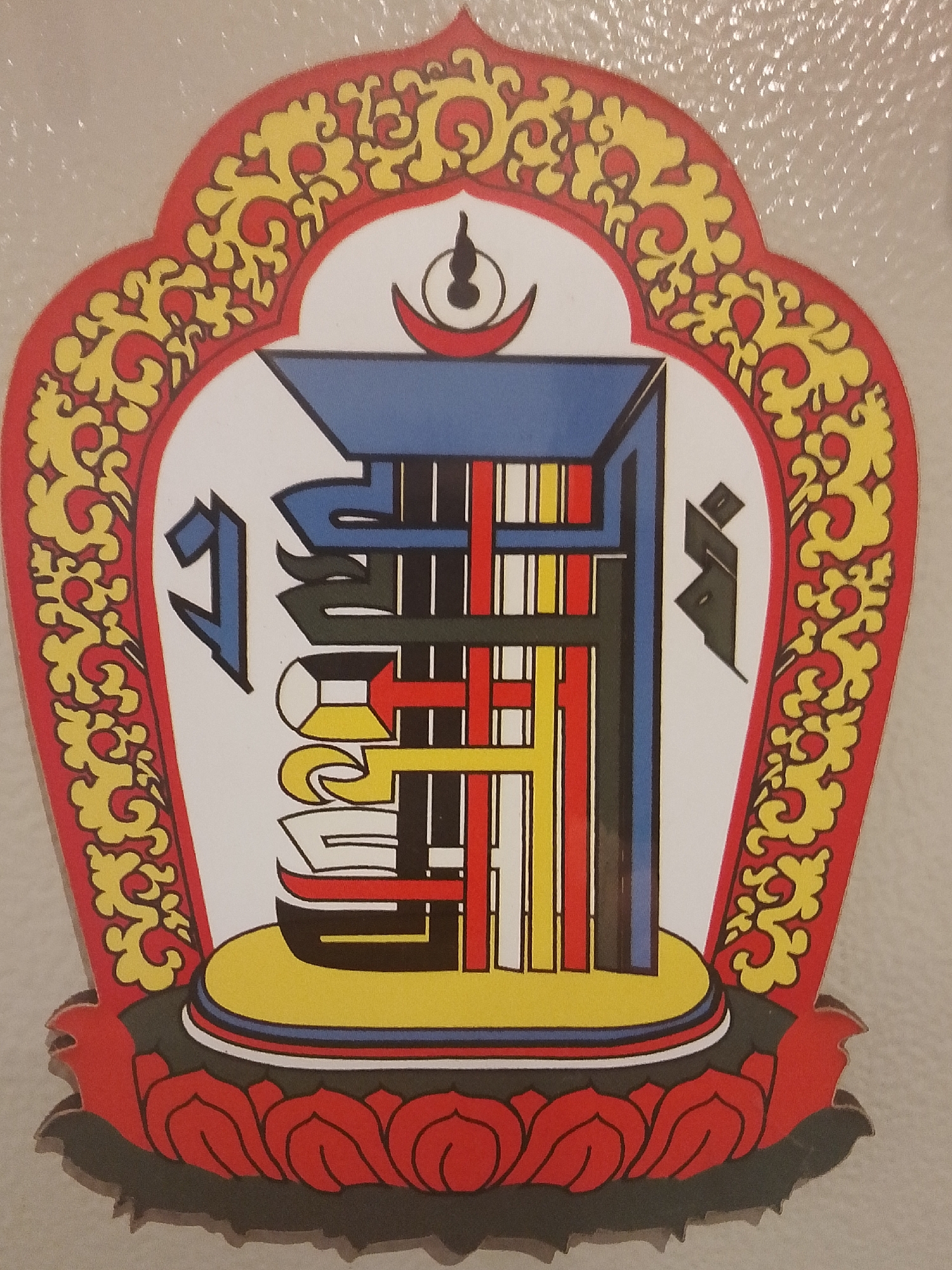 The image shows a stylized emblem with a red and yellow floral border containing a white, teardrop-shaped field. Inside the field is a multi-colored geometric symbol consisting of vertical lines in red, yellow, black, and white, intersected by horizontal lines and angular shapes in blue, black, yellow, and red. The symbol is set on a yellow platform, which rests atop a black band and a base of red lotus petals. Small, stylized symbols are present in black and blue on either side of the main geometric form. Above the geometric symbol, at the top of the field, is a crescent moon shape with a dot above it. This emblem is likely of Tibetan Buddhist origin. The location, 1-Go Maya, Russia, does not seem directly relevant or visually represented in the image.