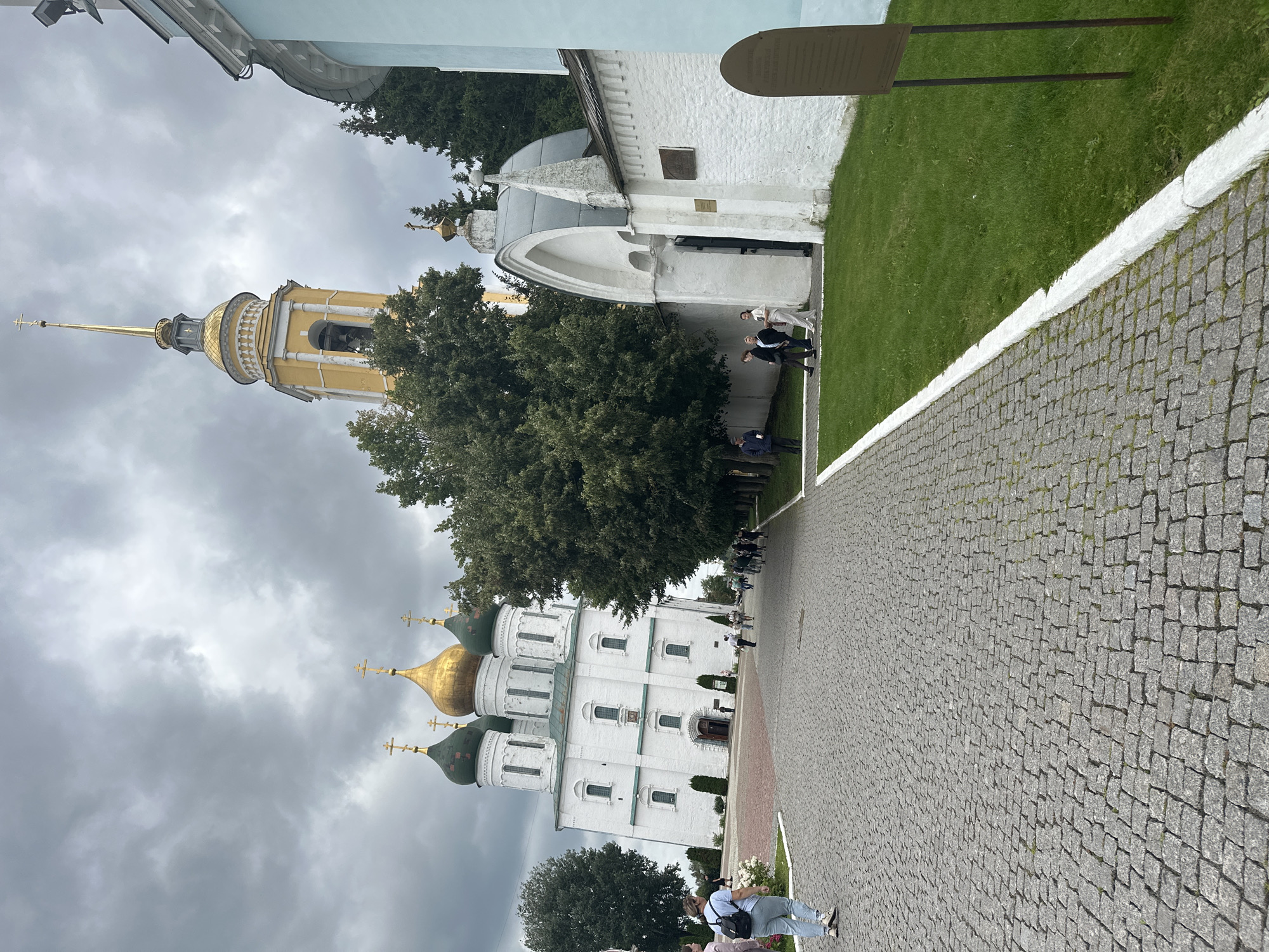 The image shows a view of the Kolomna Kremlin in Kolomna, Russia. The foreground is a cobblestone pathway leading towards a white building with green and gold domes, which is likely a cathedral or church. To the right of the pathway, there is a section of manicured green grass bordered by a white-painted curb. A tall, yellow bell tower with a gold spire stands prominently behind the white building. Several people are walking along the pathway and near the entrance to the white building. The sky is overcast with dramatic grey clouds. The overall scene depicts a religious complex with architectural elements characteristic of Russian Orthodox churches.