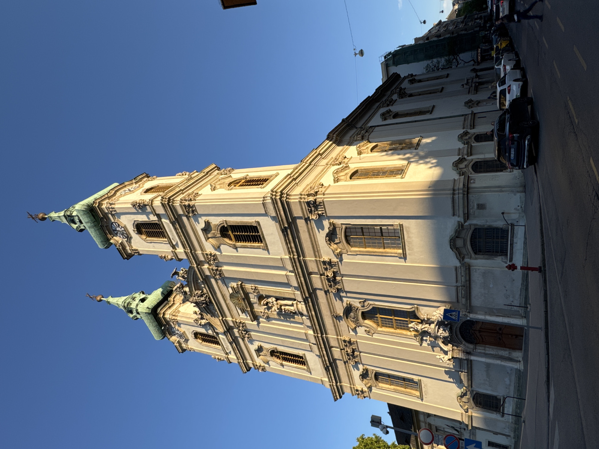 This image captures a grand Baroque-style church, its ornate facade illuminated by the warm, low-angle light of either late afternoon or early morning. The church features a light-colored exterior, rich architectural details including multiple pilasters, cornices, and decorative carvings, and numerous windows, some arched, fitted with golden-hued grilles. Two prominent towers rise from the structure, each topped with a distinctive green, pointed spire crowned with a finial. A clock face is visible on the upper part of the right tower. Statues are placed within niches on the building's facade.
The church occupies the left side of the frame, standing alongside a paved urban street on its right. The sky overhead is clear and a vibrant blue. Along the street, several cars are parked in a row, with a dark SUV prominent in the mid-ground. One person, dressed in dark attire, is visible walking further down the street.
Street furniture includes a bright red fire hydrant, various street signs—a blue pedestrian crossing sign, a circular blue sign with a red diagonal line, and another sign indicating an arrow and "10 m"—and overhead utility wires. Parts of the street and the lower sections of the church are cast in shadow due to the sun's position, enhancing the three-dimensional quality of the architecture. The scene depicts a historic building within an urban environment during a serene time of day.