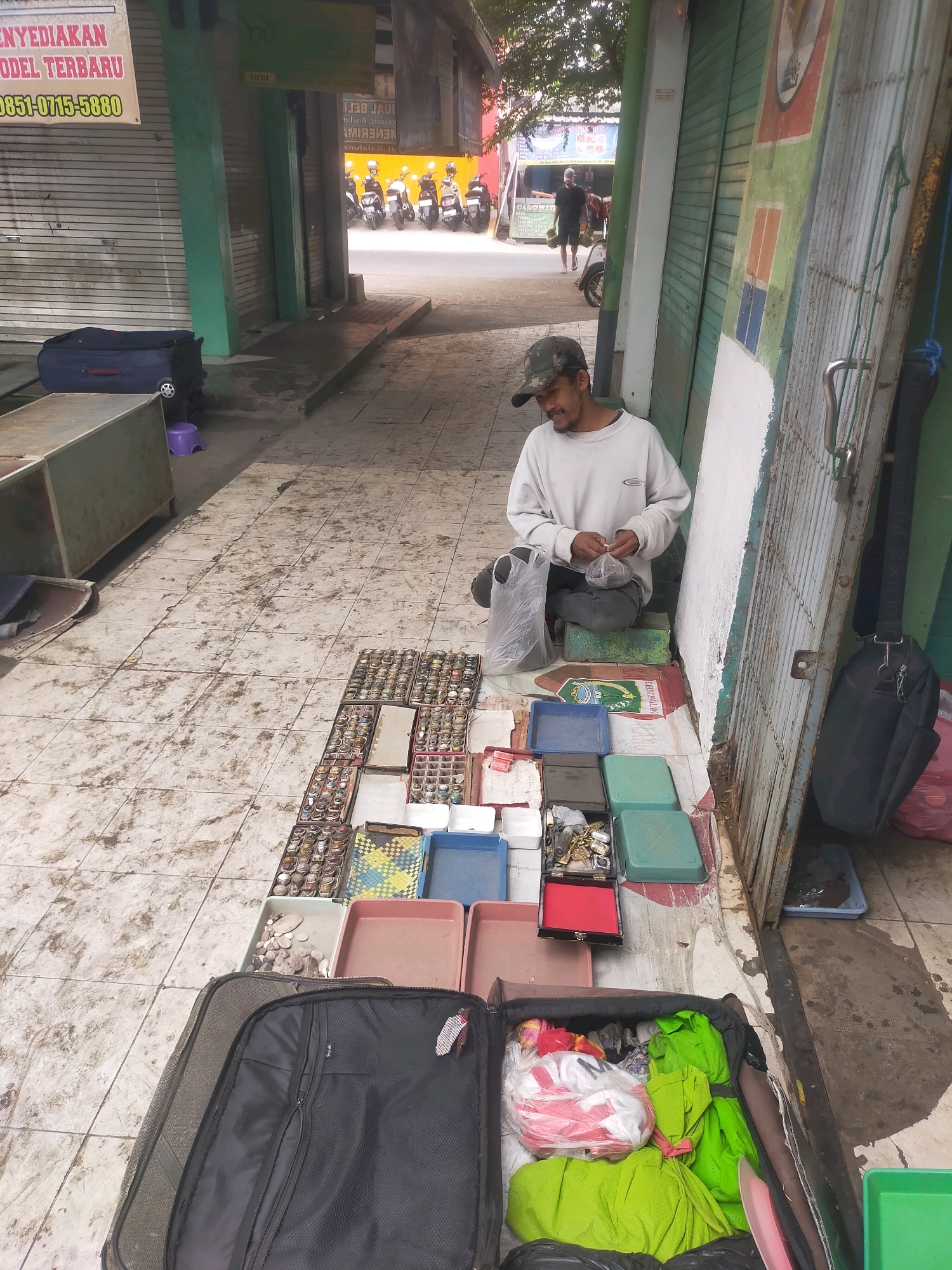 In the city of Gondanglegi, Indonesia, a man wearing a camouflage baseball cap and a light-colored long-sleeved shirt sits on the ground. He is positioned in a narrow outdoor marketplace, adjacent to a building with closed metal roll-up doors. In front of him, he has laid out a diverse collection of items for sale on the tiled ground. These include numerous trays filled with what appear to be rings, as well as other assorted jewelry and trinkets. Scattered amongst the jewelry are several colorful plastic containers. To his right, a black duffel bag hangs from a metal door frame. To his left, a large open suitcase is filled with various items, including bundles of bright green fabric and plastic-wrapped packages. The ground in the foreground is tiled and shows signs of wear and some discoloration. In the background, more roll-up doors and a street with parked motorcycles are visible, suggesting a busy commercial area.