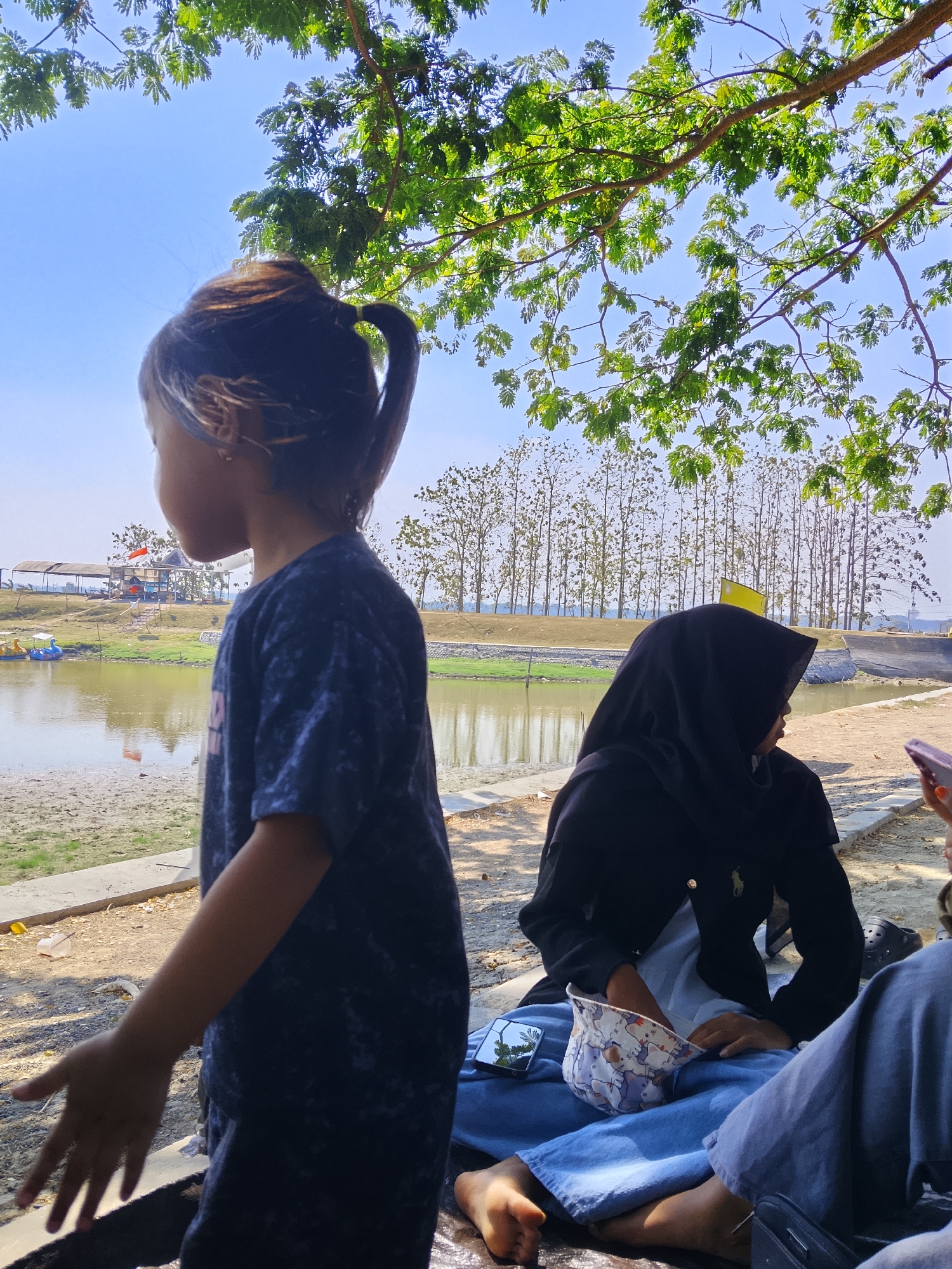 The image depicts an outdoor scene with a body of water in the background, likely a lake or pond. In the foreground, two individuals are present. A young girl, on the left, is looking away from the camera, wearing a dark blue t-shirt. Her hair is pulled back in a ponytail. To her right, a person wearing a black hijab and a black jacket sits on the ground, facing away from the camera and towards the water. This person is also wearing blue pants. Scattered around them are a few personal items, including a hat with a pattern of elephants, a smartphone, and what appears to be a pair of shoes. In the background, there are trees and a structure with a red flag, possibly part of a recreational area. The overall setting suggests a park or a lakeside recreational spot, potentially within or near the city of Ngadrin, Indonesia, given the context. The sky is clear and blue, indicating a sunny day.