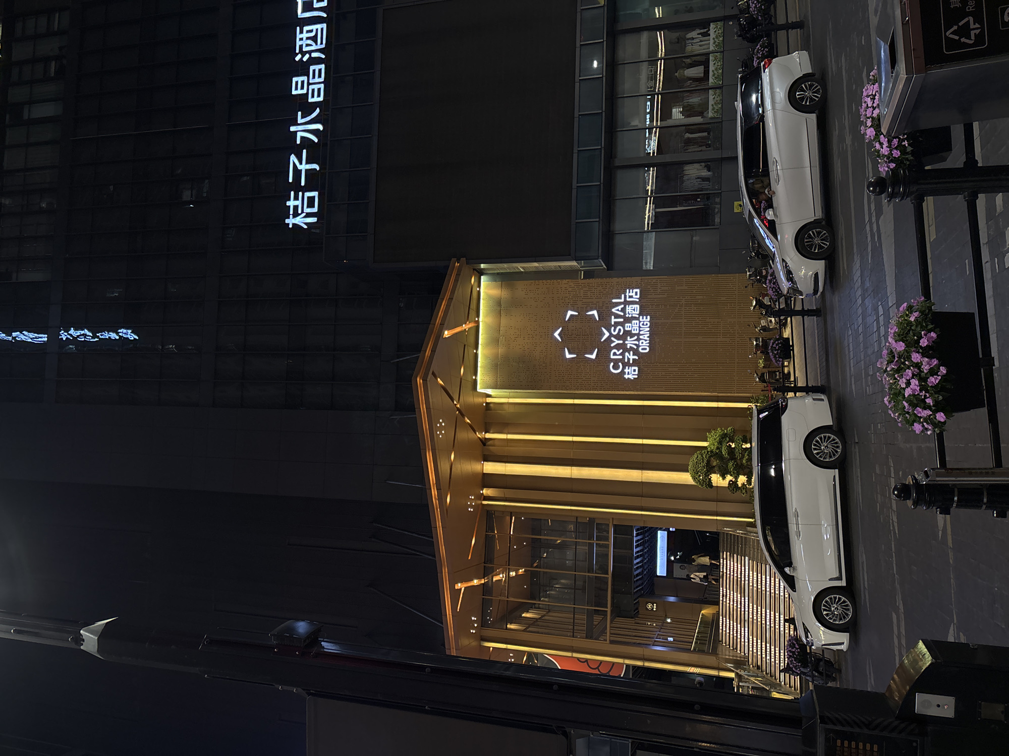 The image shows the exterior of the Crystal Orange Hotel in Yuzhong District, China, at night. The hotel entrance features brightly lit vertical panels and a sign with the hotel's name in both English and Chinese. A taller building with Chinese characters illuminated vertically is visible to the left of the hotel. Two white cars are parked along the street to the right of the hotel entrance. Flower planters line the sidewalk. Streetlights and building architecture dominate the foreground and background. There are people by the hotel's stairs.